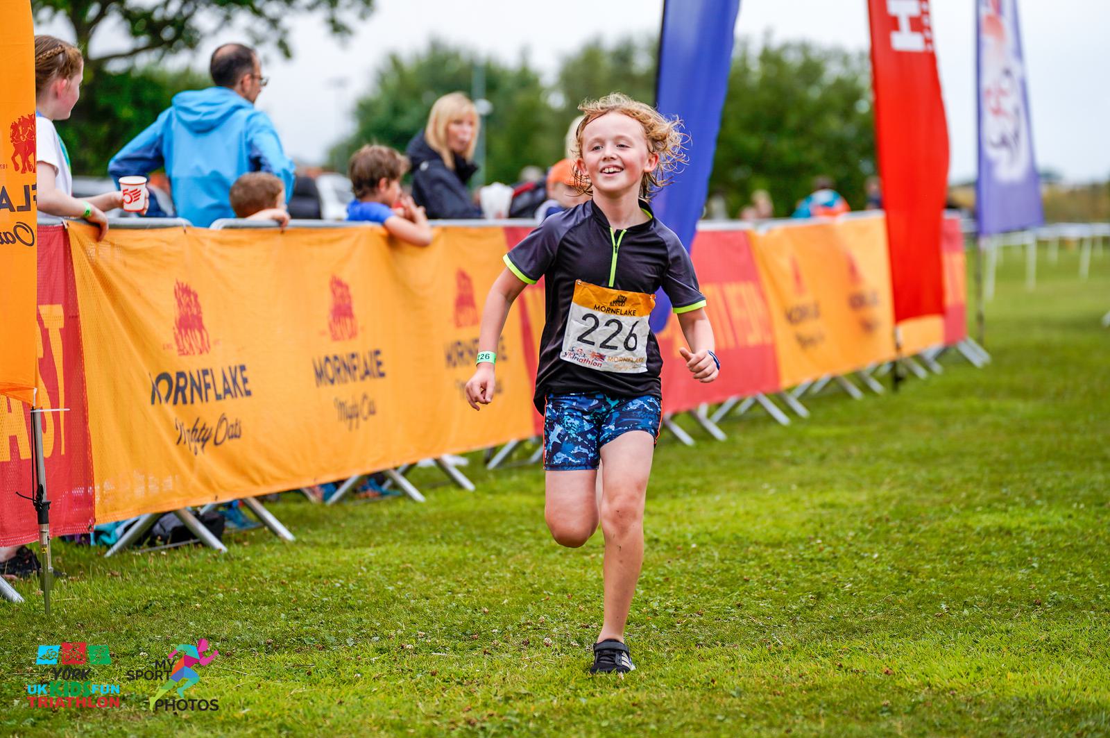 A young child with curly hair runs joyfully towards the finish line of a race, wearing a black and blue outfit and numbered bib 226. Spectators behind orange barriers cheer on the participants. The background features sponsorship banners and a somewhat overcast sky.