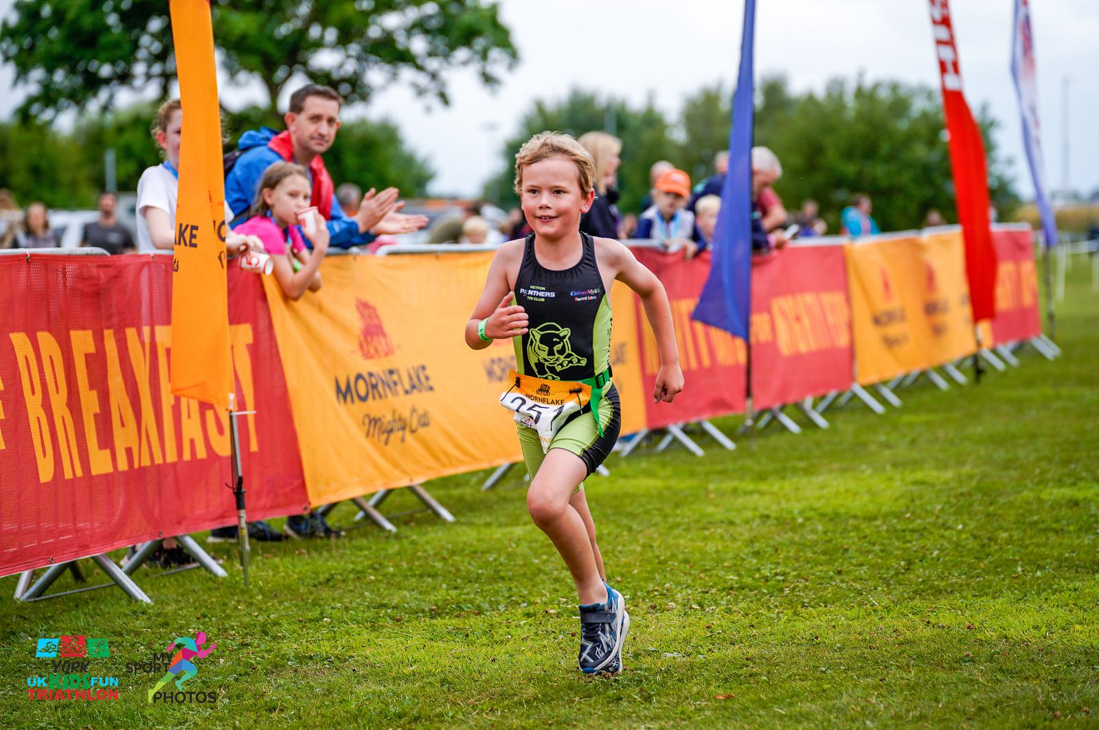 A young child wearing sports gear runs on a green field during a triathlon event. Spectators line the course behind a bright orange barrier with promotional text. Event banners and flags are visible in the background, and photographers are capturing the moment.