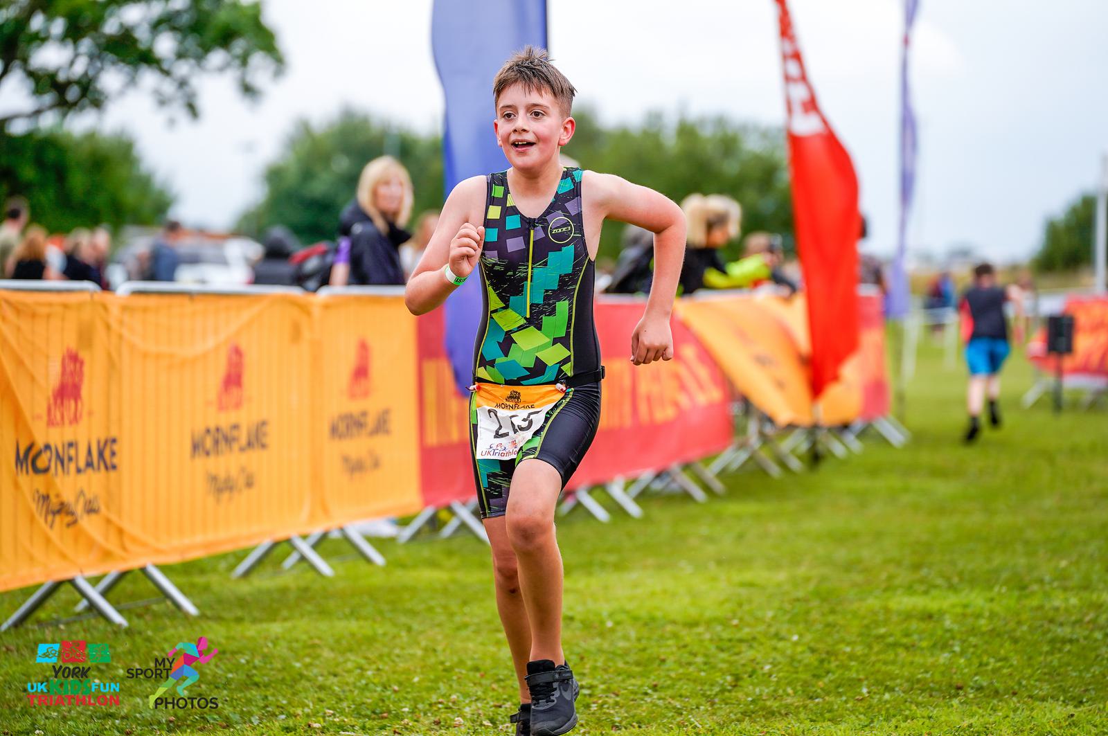 A young athlete, dressed in a green and black triathlon suit, runs toward the finish line of a race. He is wearing the bib number 257 and looks determined. Spectators and colorful banners line both sides of the course, and other participants are visible in the background.