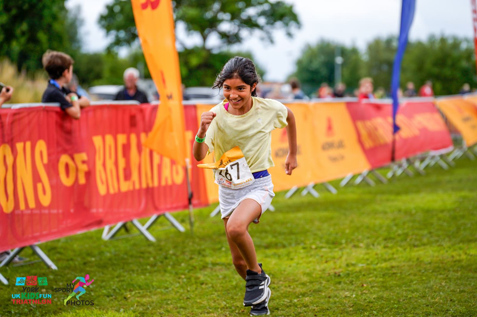 A young girl wearing a yellow shirt and white shorts is running towards the finish line of a race with a joyful expression. She holds a small flag and wears a bib number 67. The background shows colorful banners and other participants in a green outdoor setting.