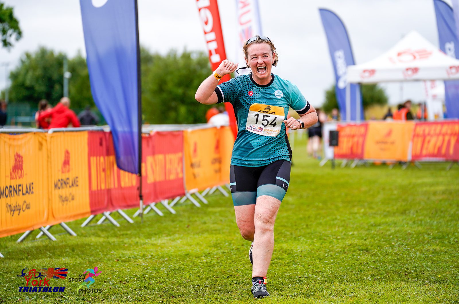 A joyful athlete wearing a green and black outfit runs towards the finish line, raising a fist in celebration. She has bib number 154 and the background shows event branding and a green field under a cloudy sky.