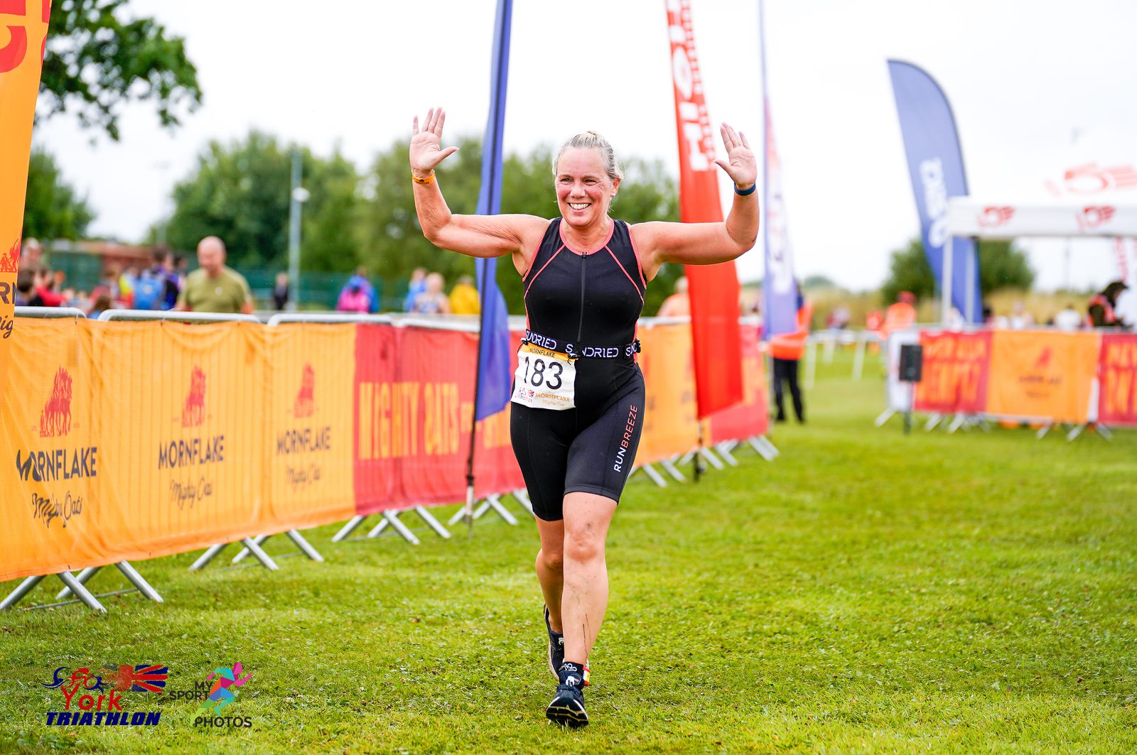 A woman wearing a black triathlon suit, numbered 183, is running with both arms raised in celebration towards the finish line of a triathlon event. Brightly colored banners and spectators line the course in the background on a grassy field.
