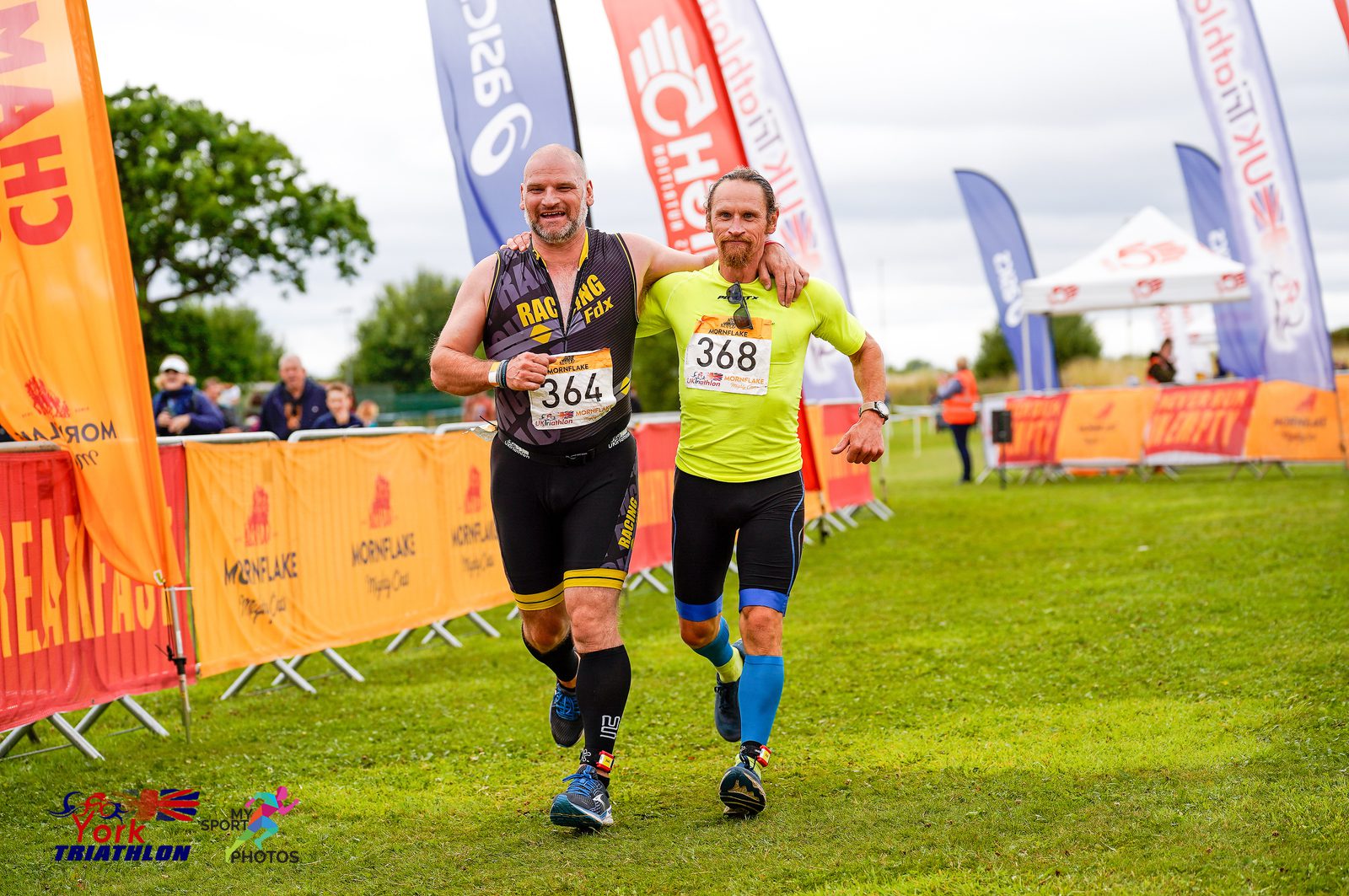 Two athletes, one wearing a black tri-suit and the other in a neon yellow top, cross the finish line together at a triathlon event. They appear joyful and exhausted. The finish line is adorned with colorful banners and flags, and spectators stand in the background.