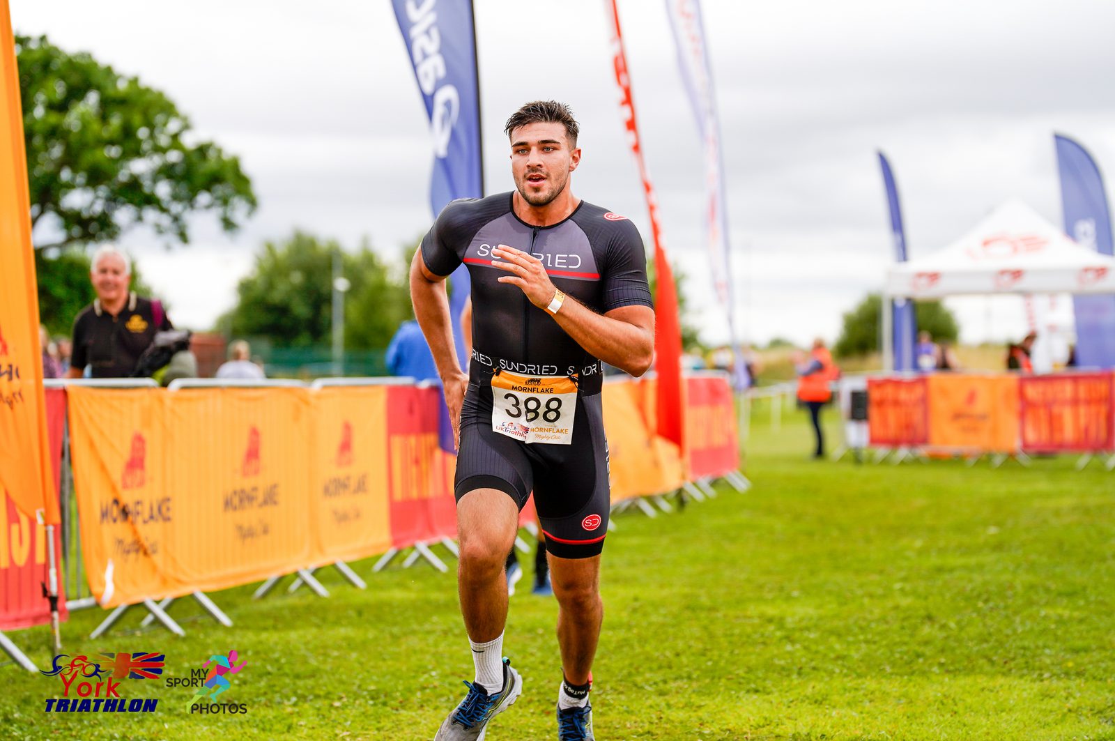 A male athlete in a racing bib numbered 388 sprints towards the finish line at a triathlon event. He wears a black and red athletic suit and white sneakers. Event banners and spectators are visible in the background on a grassy field.