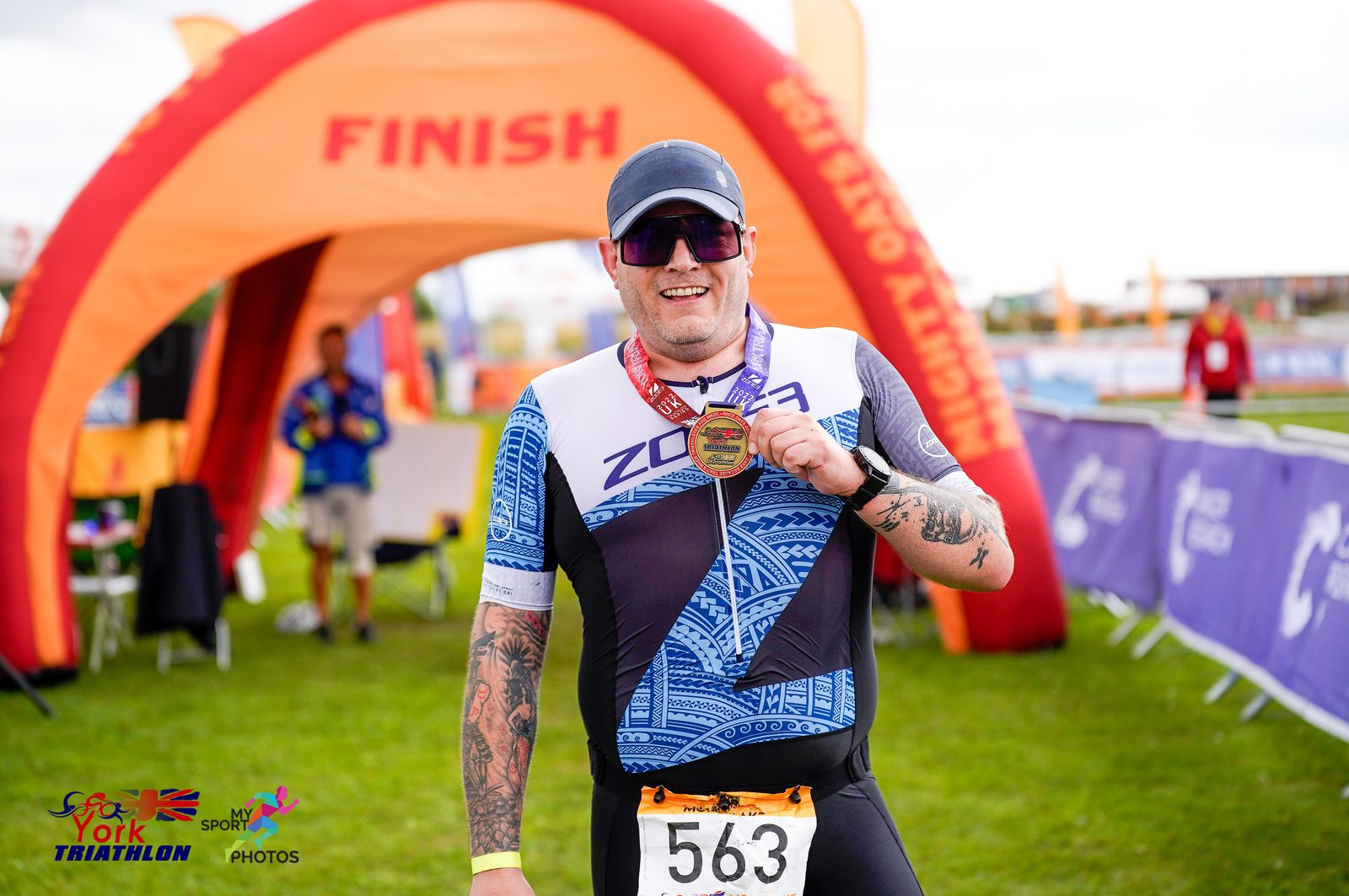 A person wearing sunglasses and a cap proudly holds up a medal while standing in front of a finish line at a triathlon event. They are wearing a sports outfit with the number 563. The background includes event banners and an inflatable archway with "finish" written on it.