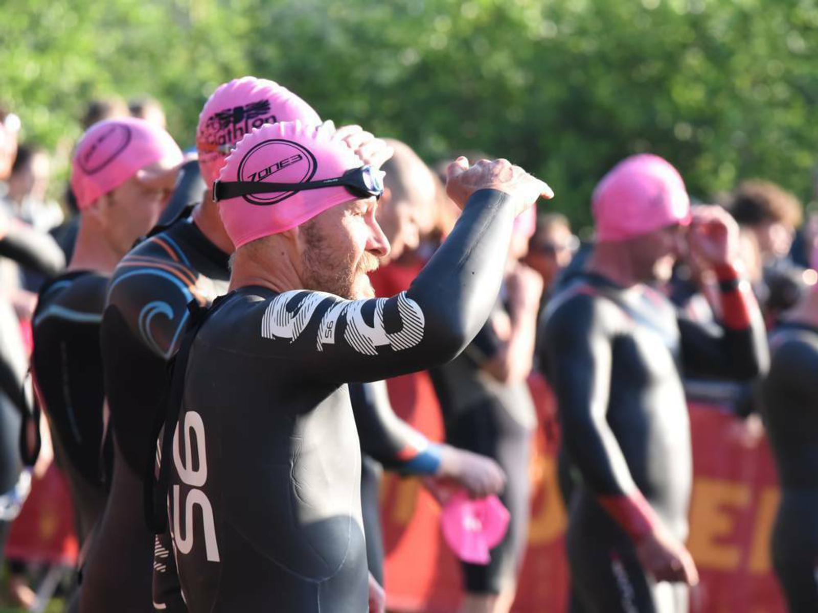 A group of triathletes wearing black wetsuits and pink swim caps stand in a line, preparing for the swimming segment of a race. One participant in the foreground adjusts his goggles while people behind him appear to be chatting and waiting. Trees are visible in the background.