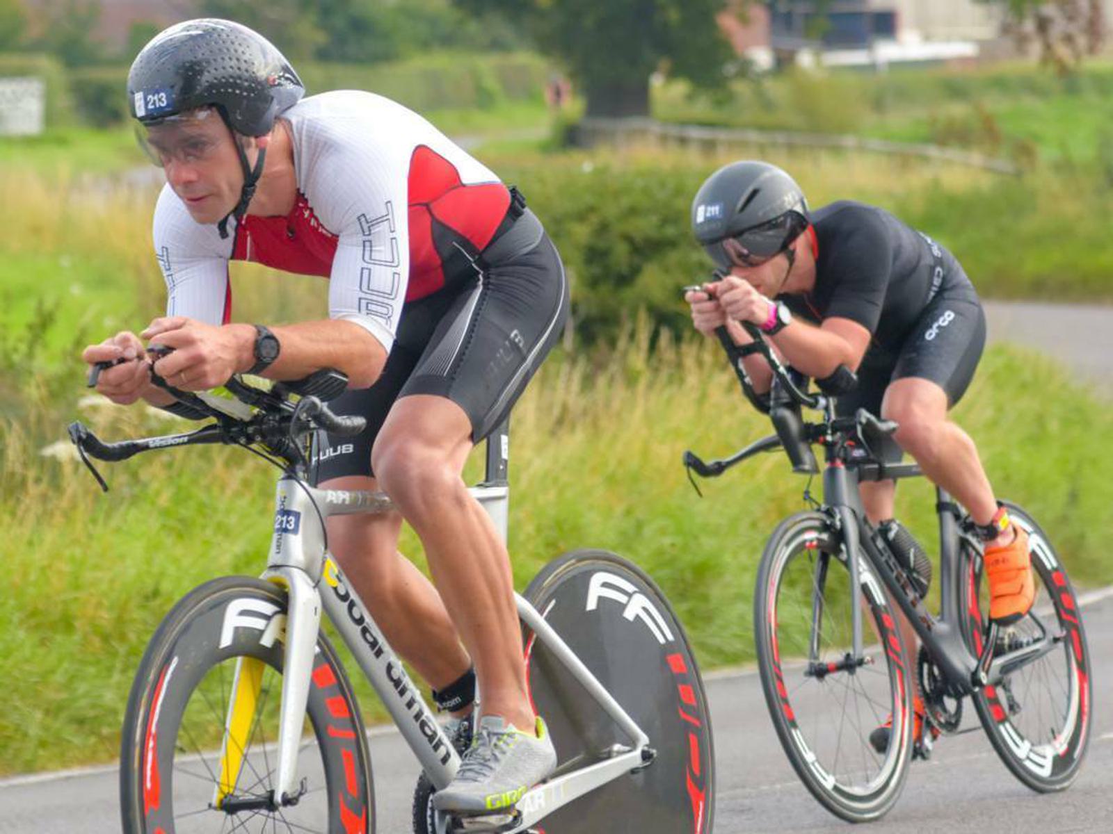 Two cyclists, wearing aerodynamic helmets and cycling gear, ride high-performance bikes on a road in a race. The cyclist in the foreground wears a white and red outfit, while the one behind him wears all black. They are focused and in racing positions.