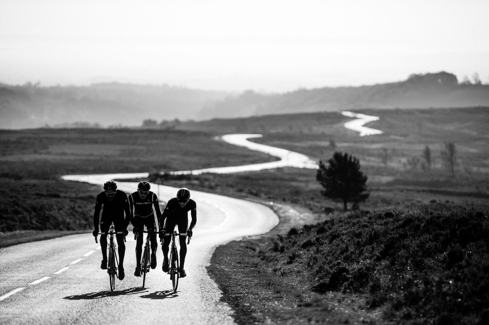 Black and white image of three cyclists riding along a winding road in a vast, open landscape. The road meanders through rolling hills under an expansive, brightened sky. The cyclists are dressed in similar dark gear, riding closely together.