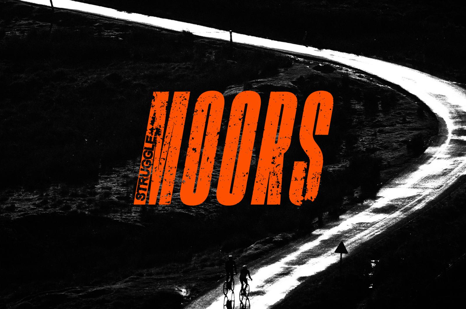 A dark, high-contrast image shows two cyclists on a winding, wet road at night. The text "STRUGGLES MOORS" in large, bold, distressed orange font is superimposed over the image. The background is shadowy, with the bright road creating a striking visual path.