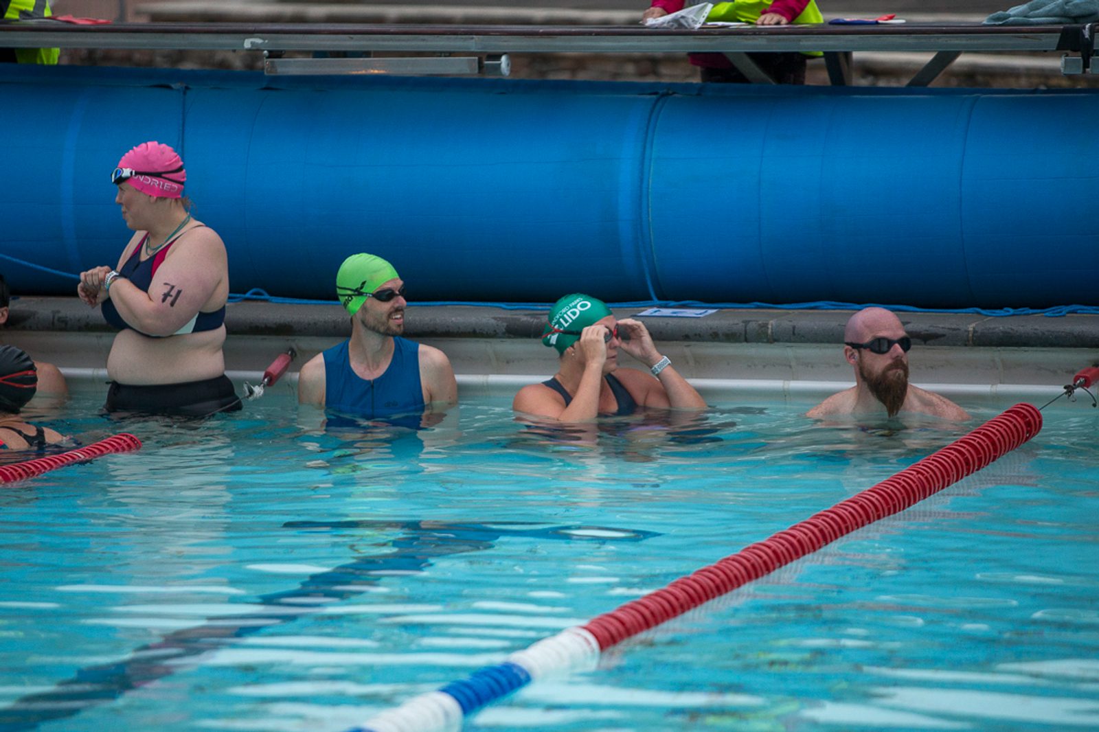 A group of swimmers in a pool lane, wearing swim caps and goggles. Four people are visible: two are adjusting their goggles, one is standing out of the water with a pink swim cap and black swimsuit, and one is floating with a beard. Lane dividers are visible.