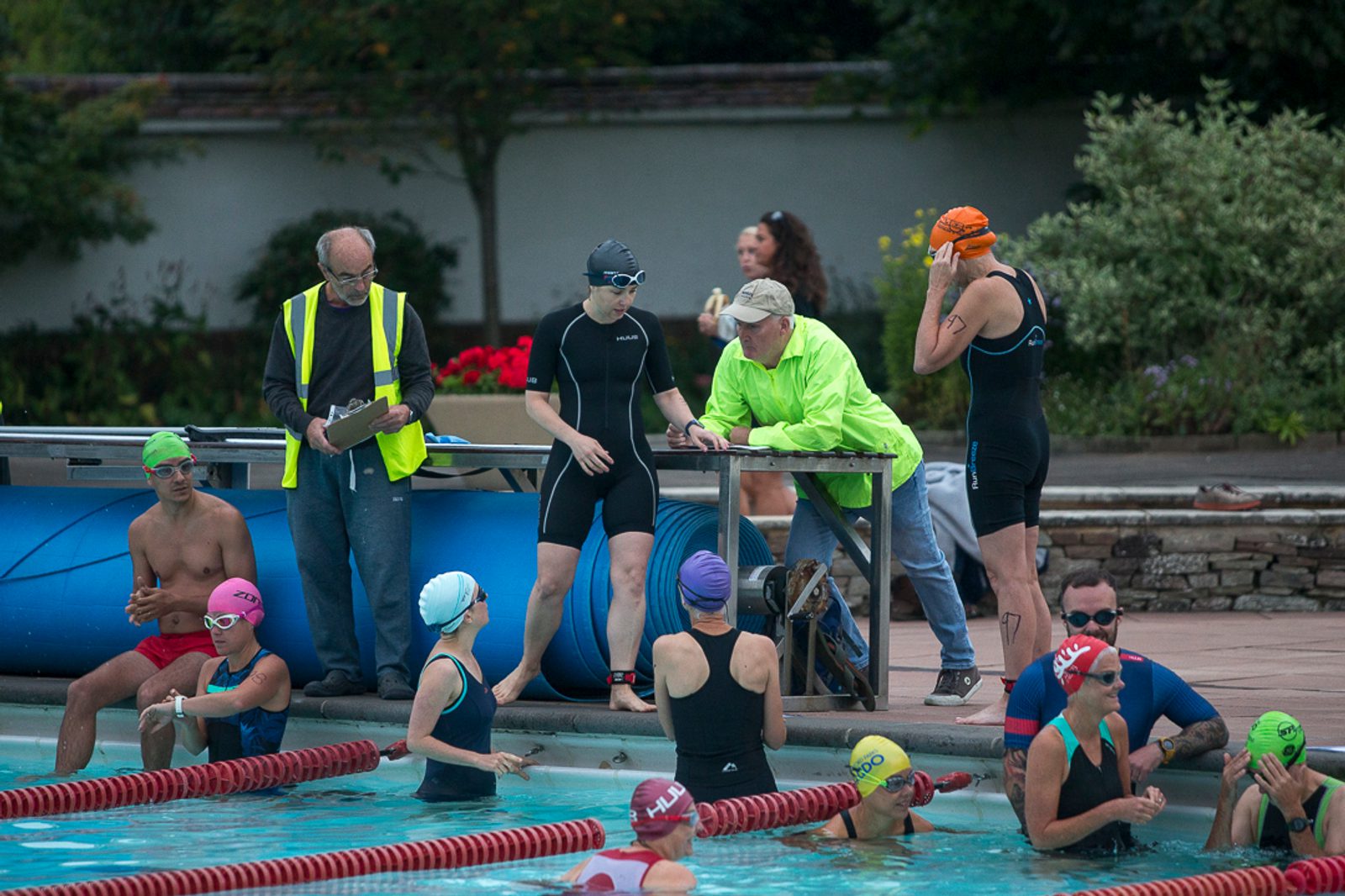 Several swimmers, some wearing brightly colored swim caps, are either standing or in the pool, participating in a swim event. Two officials are on deck, one in a bright yellow vest taking notes. Trees and a stone wall make up the background.