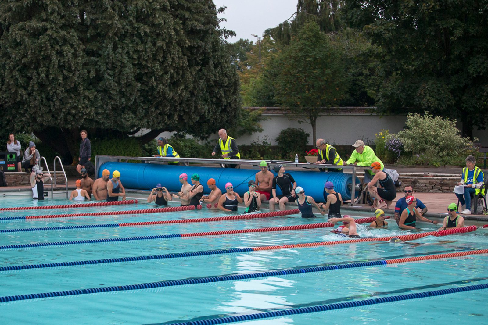 A group of swimmers in colorful swim caps and goggles sit or stand at the edge of an outdoor swimming pool, preparing for a race. Several people in yellow high-visibility vests line the poolside, officiating or providing support. There are lush trees and foliage in the background.