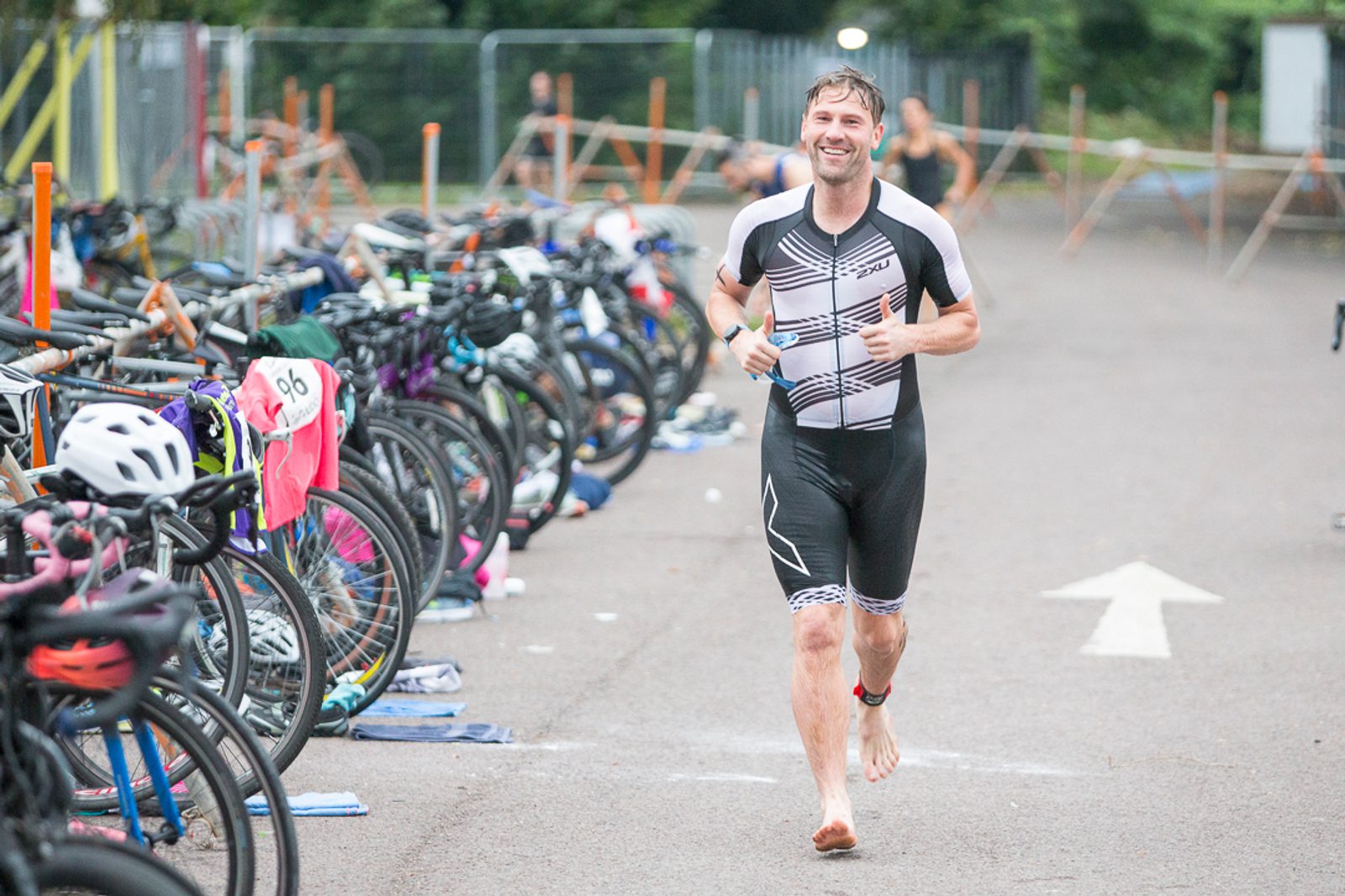 A man is running barefoot through a transition area during a triathlon. He's wearing a black and white triathlon suit and smiling. Bicycles are parked on racks to the left, with various gear laid out on the ground. The area is outdoors with greenery in the background.