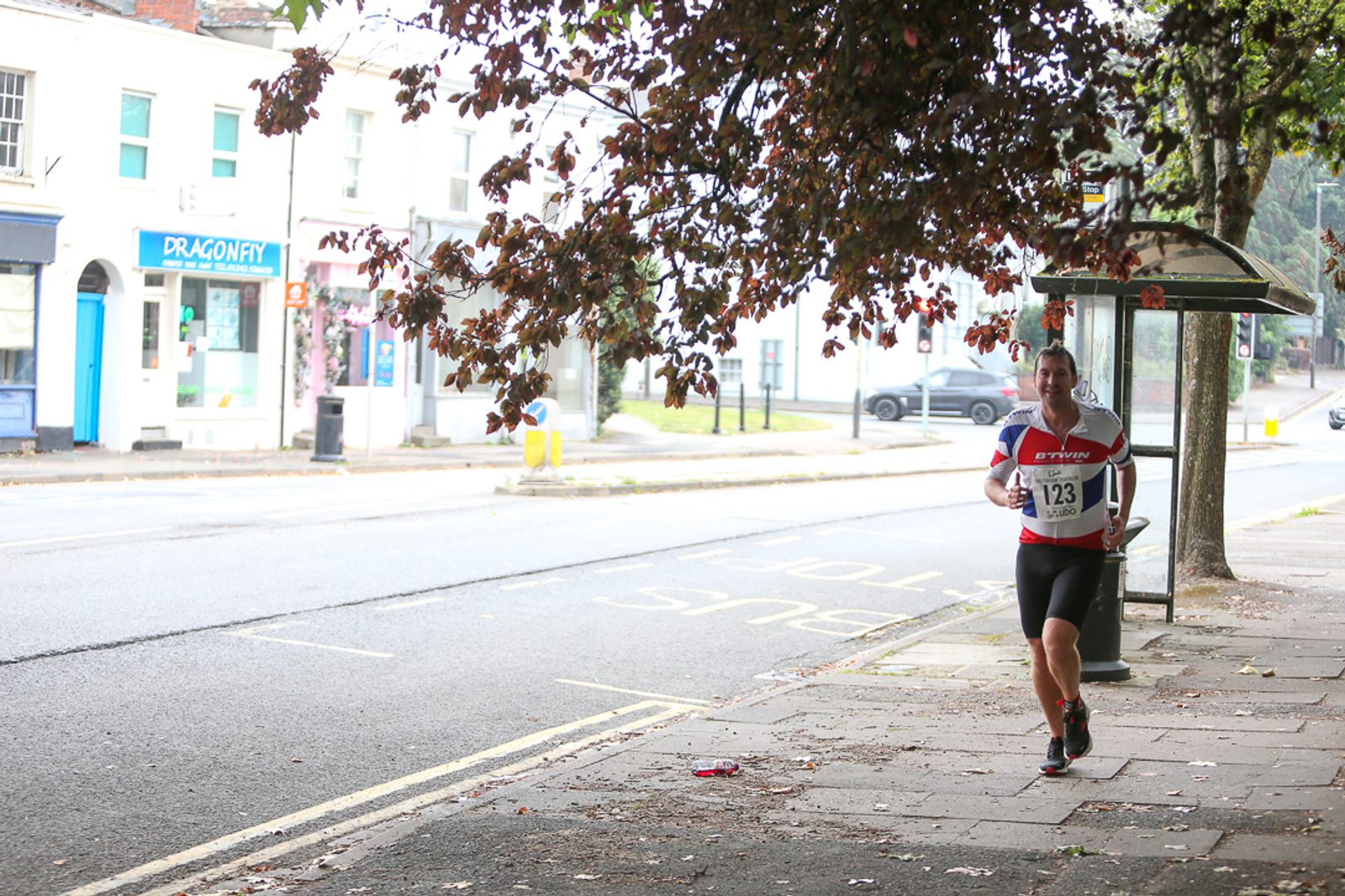 A person is jogging along a sidewalk, wearing a race bib numbered 123. The street is lined with trees and shops, including one with a "DRAGONFLY" sign. The weather appears overcast, and the runner looks focused. Fallen leaves are scattered on the sidewalk.