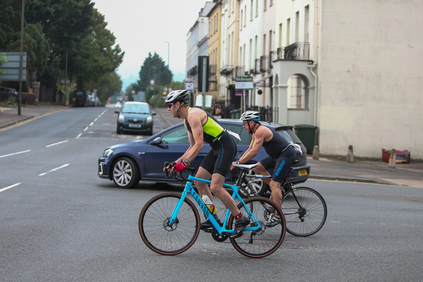 Two cyclists in sports gear ride down a city street. One cyclist on a blue bike wears a yellow and black outfit, and the other on a black bike wears a grey outfit. Cars are visible on the road and parked along the side. Buildings and trees line the street.