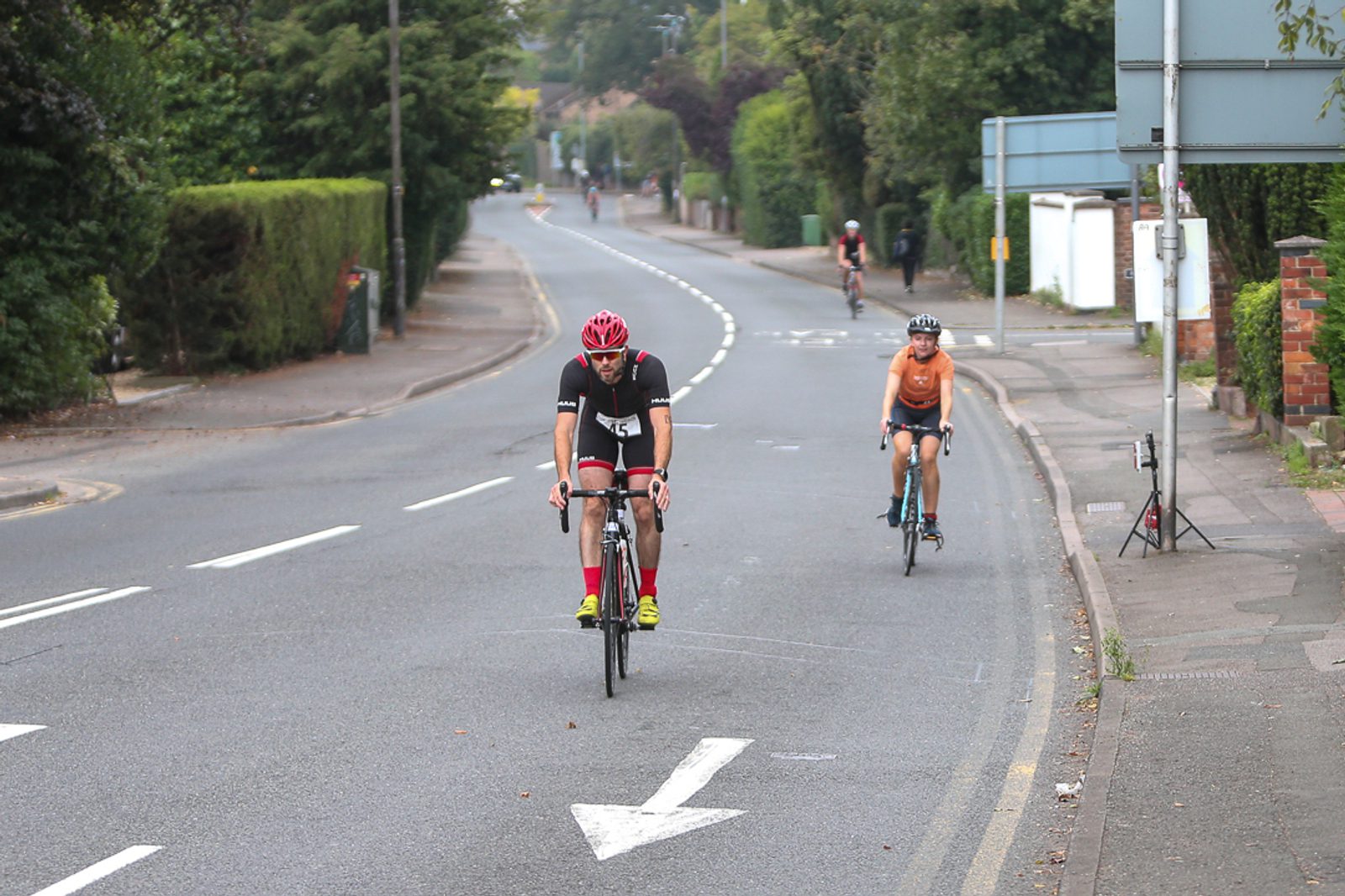 Two cyclists ride on a suburban road. The leading cyclist wears a red helmet, black kit, and yellow shoes. The second cyclist, in an orange shirt and black shorts, follows behind. The road is bordered by trees and bushes, with an arrow painted on the asphalt.