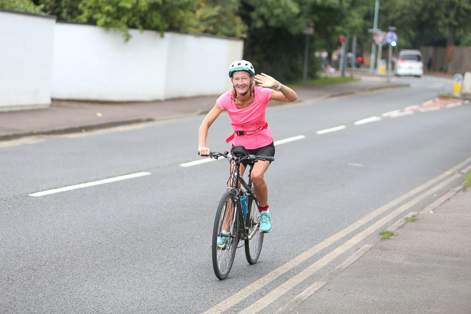 A woman in a pink shirt and helmet is riding a bicycle on a suburban street, smiling and waving with one hand. She is on a paved road with markings, and there are trees and a white wall in the background.