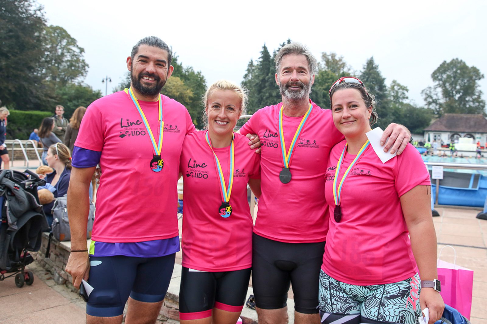 Four people wearing pink shirts and medals stand side by side smiling at an outdoor event. Trees and a pool are visible in the background, and they appear to be at a sporting event or competition.