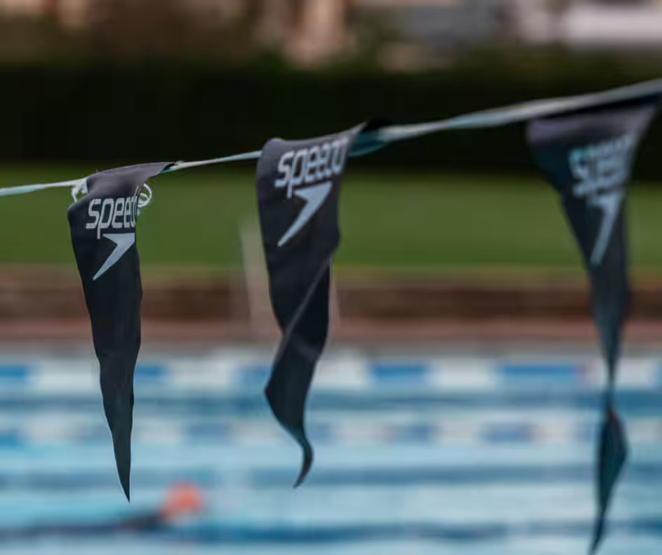 Close-up of a swimming pool lane with black Speedo flags hanging from a rope. The blurred background shows the pool's blue lanes and a swimmer, along with greenery and a building in the distance.