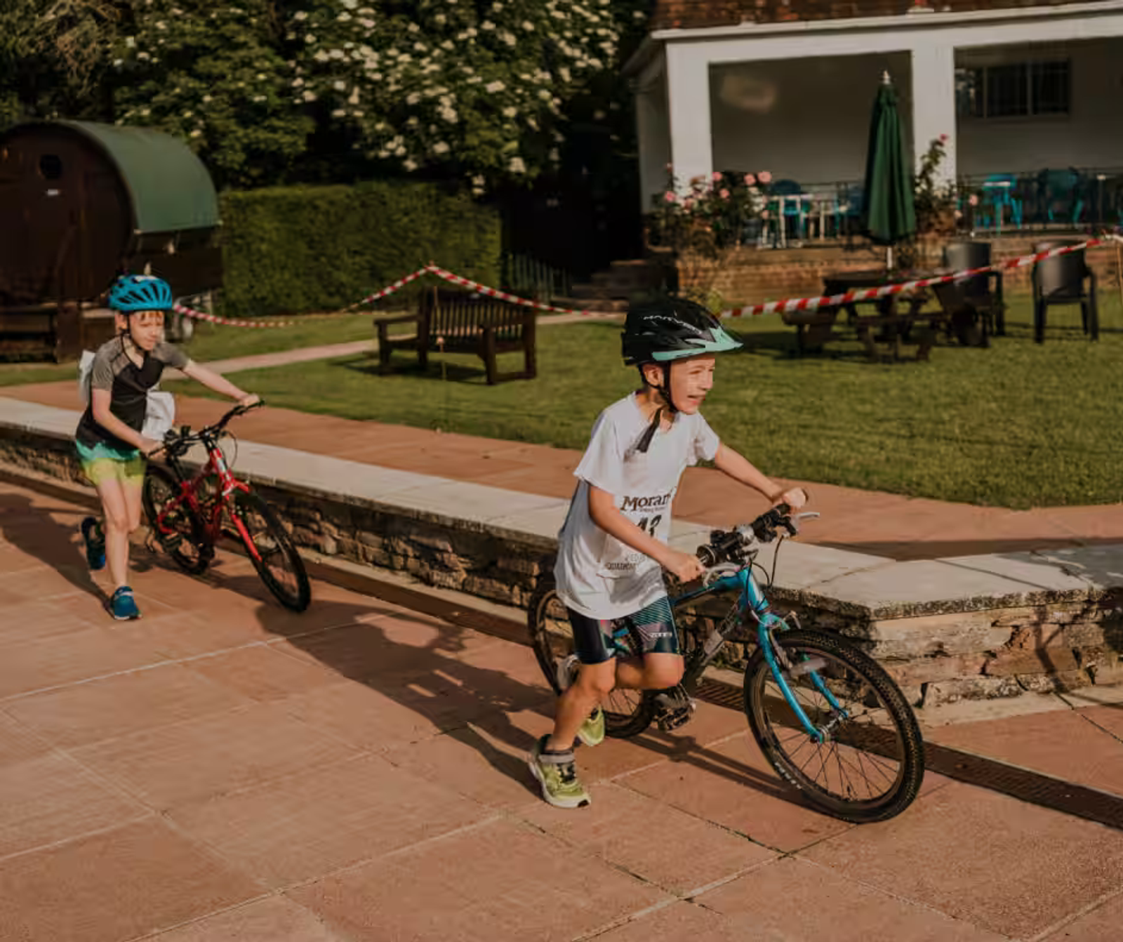 Two children wearing helmets are pushing their bicycles on a sunny path. One child is in the lead, wearing a white shirt, followed by another child in a blue shirt. They are near a building with outdoor seating and green grass in the background.