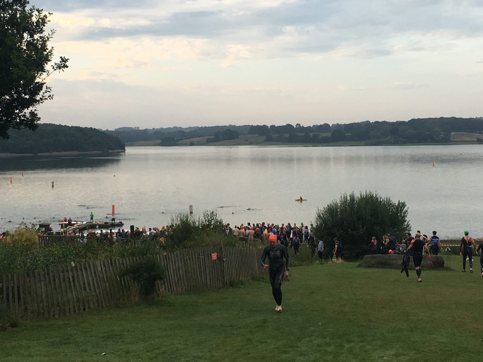 A group of people gather by the edge of a large, calm lake, some preparing to swim. The surrounding landscape features rolling hills, trees, and a partly cloudy sky. A wooden fence lines a grassy area leading to the water.