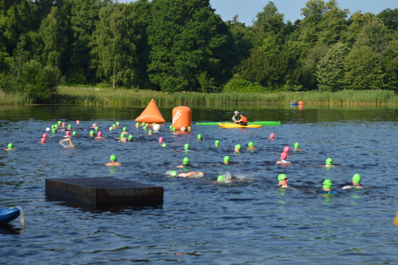 Open water swimmers in brightly colored swim caps participate in a swim race in a lake surrounded by lush greenery. Buoys mark the course, and a person in a kayak monitors the swimmers. The sky is clear and the weather appears warm.