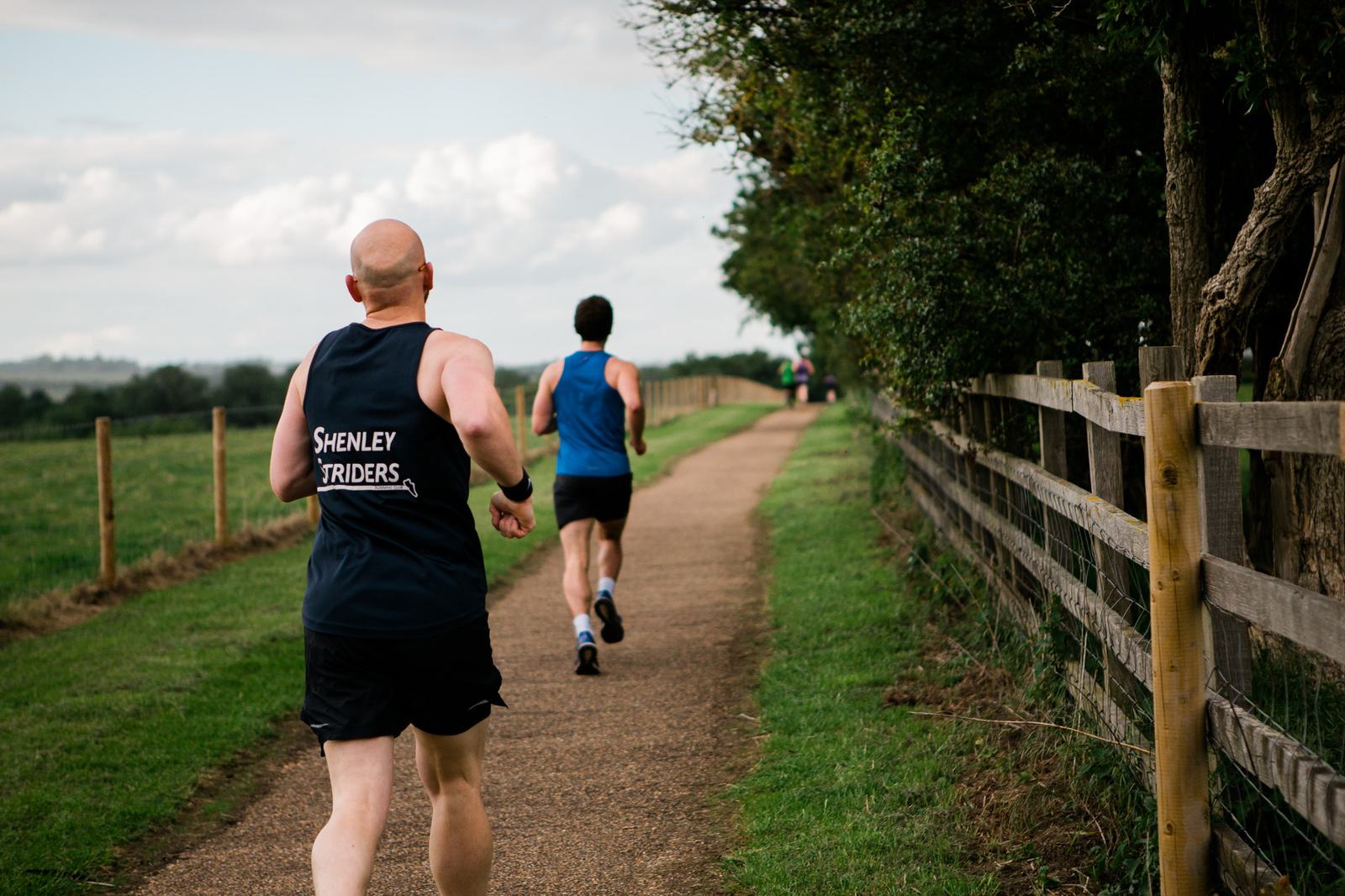 Two men are running on a path beside a wooden fence and grassy field. One wears a blue tank top with "Shenley Striders" on the back. The sky is cloudy, and trees line the path on the right.