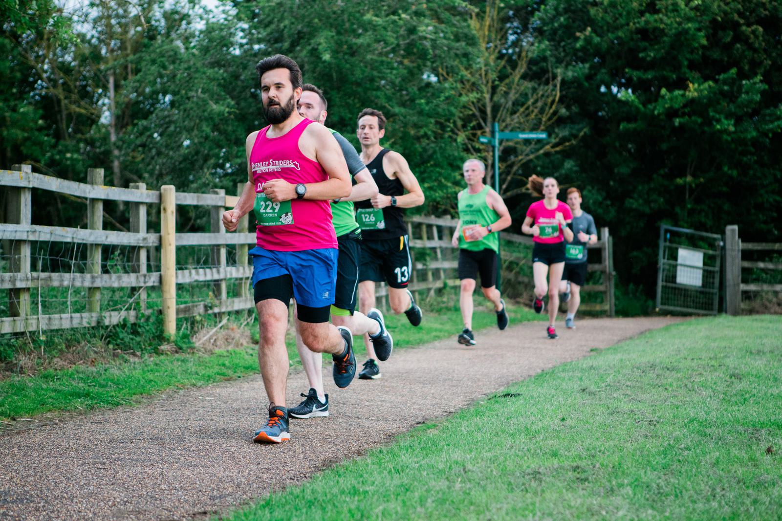 A group of runners participates in a race on a paved path surrounded by greenery. The lead runner wears a bright pink tank top and blue shorts. Other participants follow closely, dressed in various athletic gear. A wooden fence lines the path.