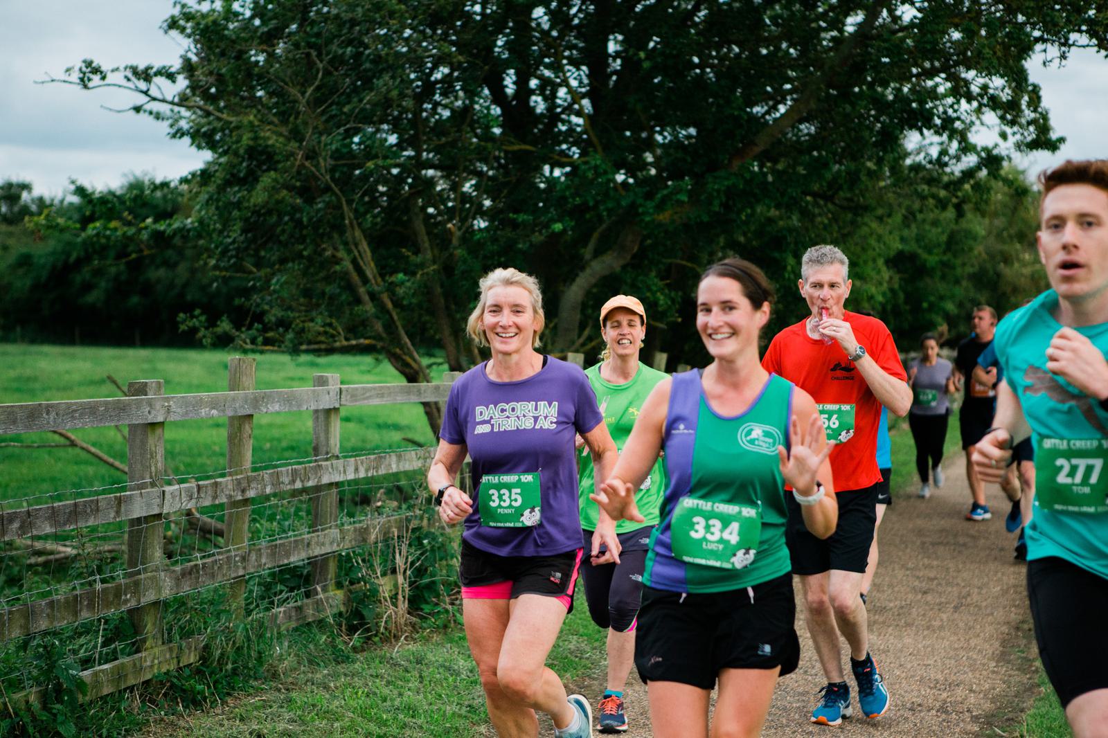 A group of runners jogs along a rural path, bordered by a wooden fence and green fields. Two women in colorful jerseys smile and wave at the camera, while others focus on their run. Trees and a cloudy sky are in the background.