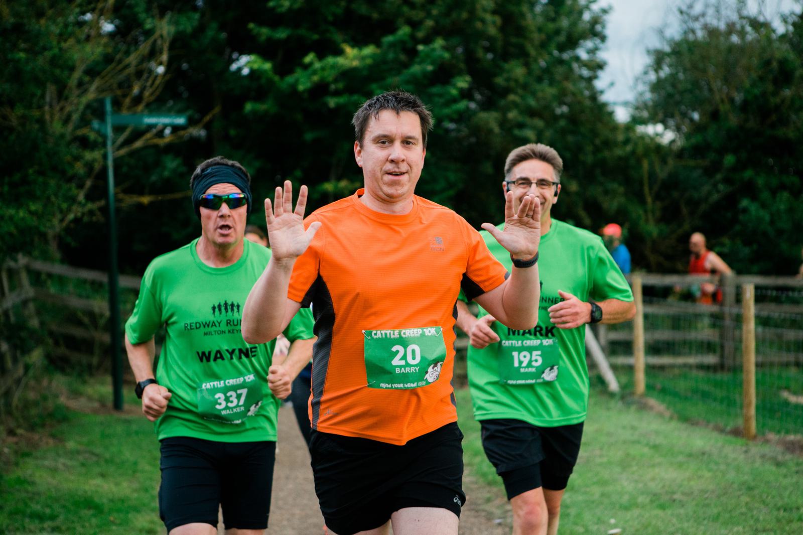Three men are jogging on a path. The man in front wears an orange shirt, smiling with hands up. Two men behind wear green shirts and sunglasses. Race bibs read "Wayne" and "Mark." Trees and a fence line the route.