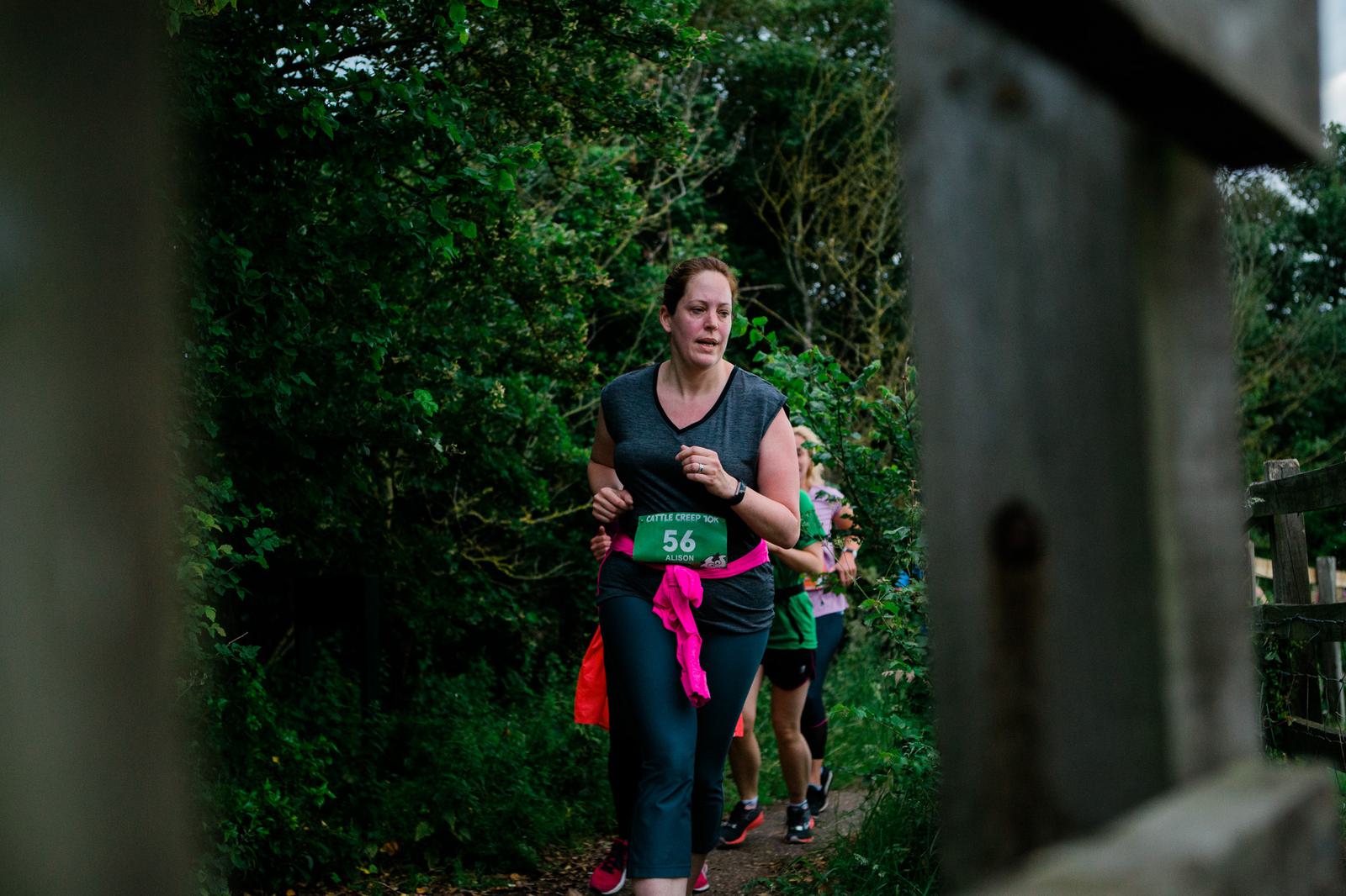 A woman wearing a race bib with the number 56 is running on a narrow, wooded path. She has a pink sweatshirt tied around her waist and appears focused. Other runners can be seen partially behind her. Trees and greenery surround the path.