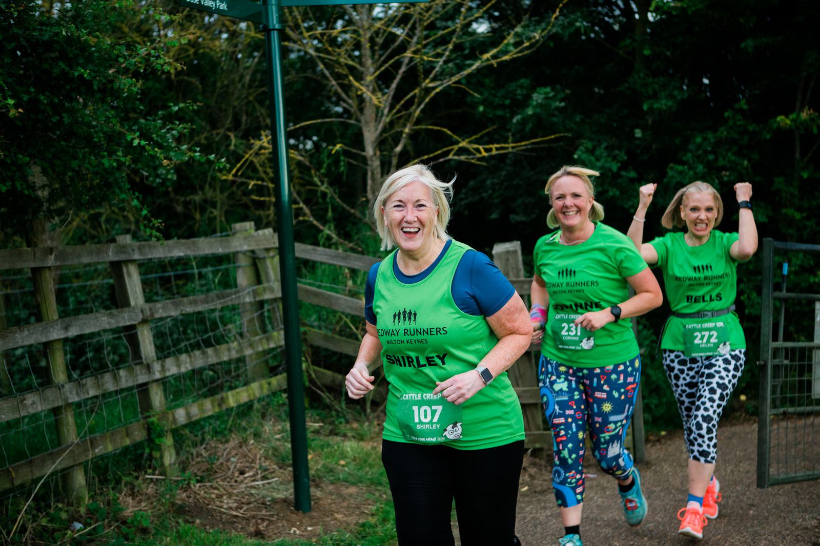 Three women wearing green shirts and running numbers participate in an outdoor race on a path lined with greenery. The woman in front smiles, followed by two more, with one raising her fist in excitement.
