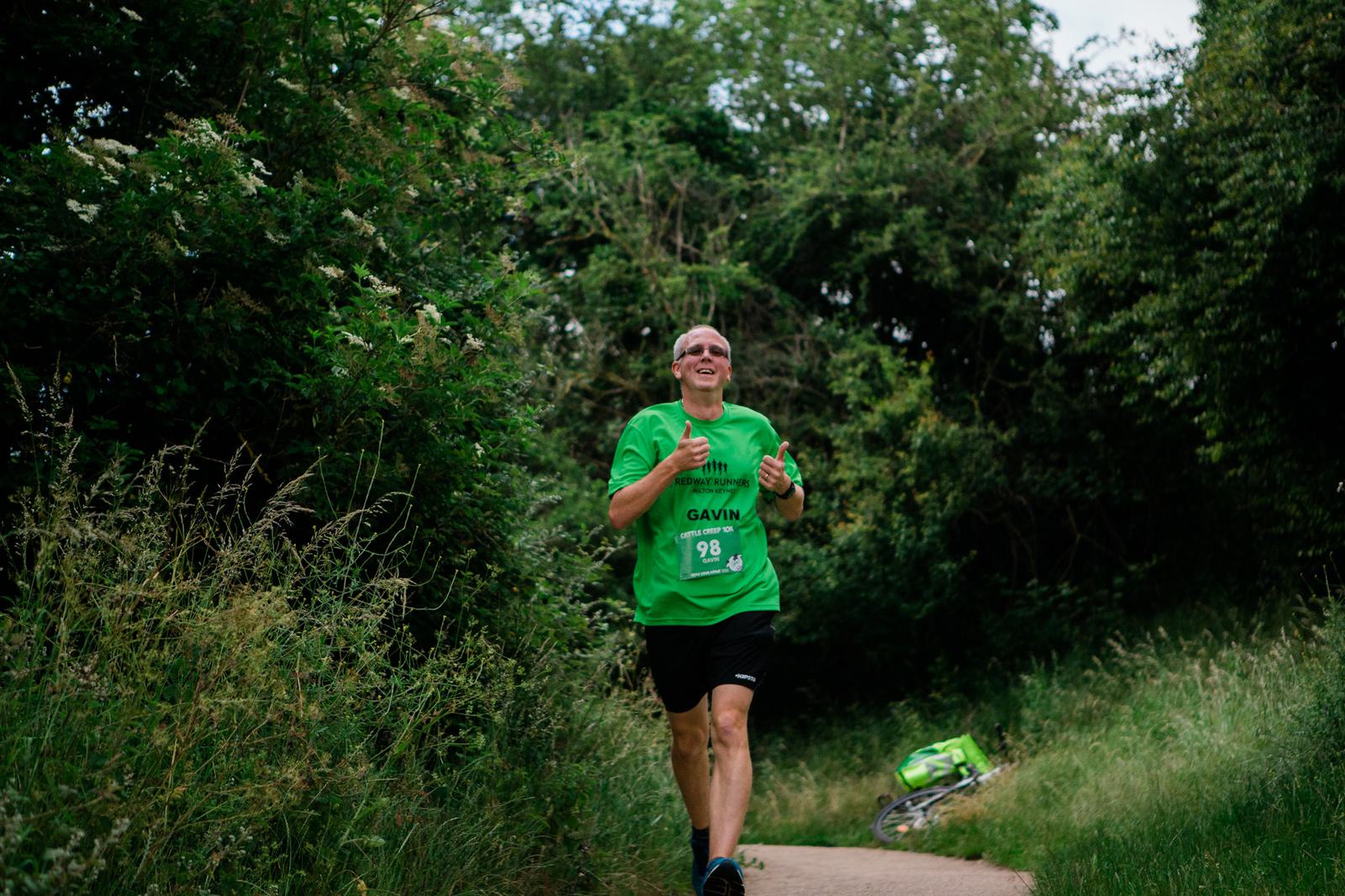 A man in a green shirt and black shorts is jogging on a path through lush greenery. He is smiling, with his hands raised in a running posture. A bicycle is partially visible on the grass beside the path.