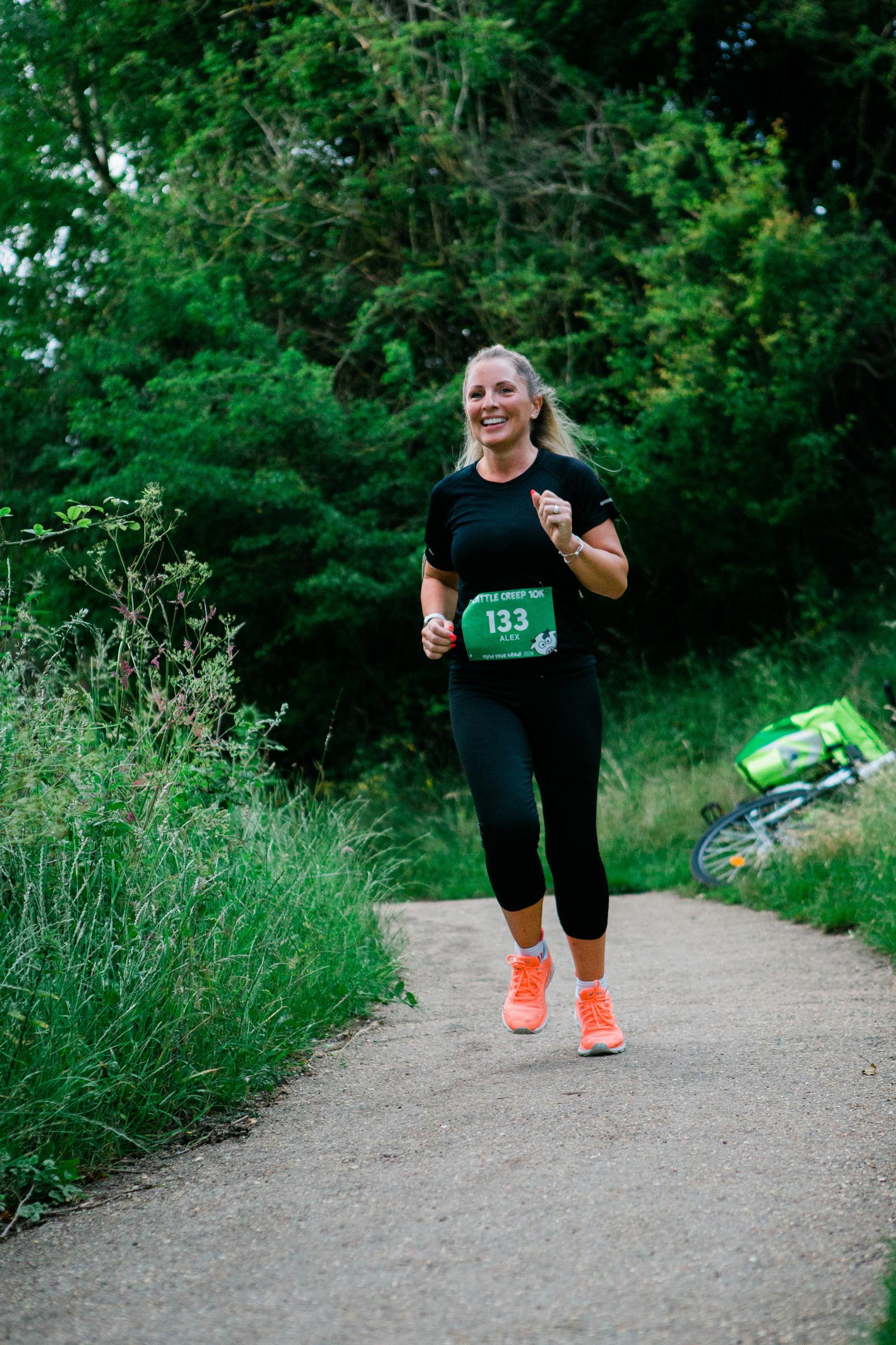 A woman jogging on a path through a green park, wearing a black outfit, orange shoes, and a race bib numbered 133. She is smiling, with trees and a bicycle lying on the grass in the background.