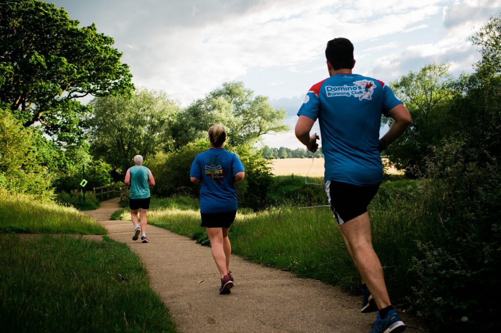 Three people jog down a winding path through a lush, green park. The sun shines brightly, casting shadows from the trees lining the path. The runners wear athletic clothing, including T-shirts, shorts, and sneakers, and the scenery is vibrant and inviting.