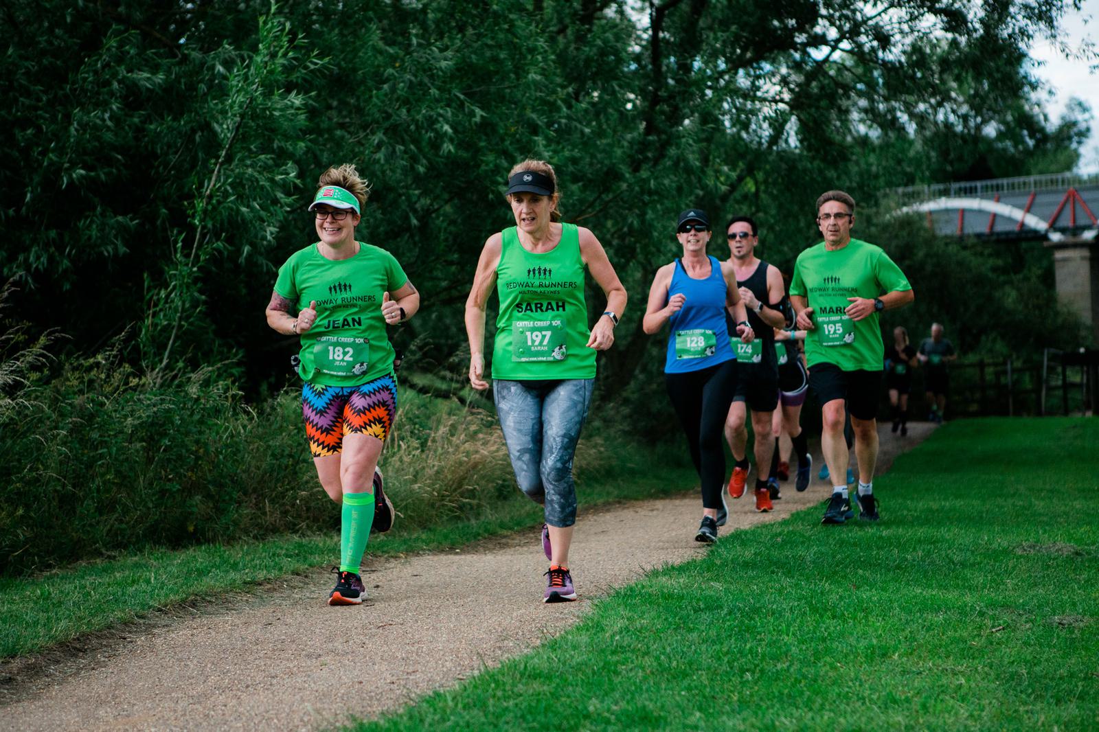 A group of runners jogs along a path in a park. They are wearing bright green shirts with numbers, indicating they're in a race or marathon. Trees and grass surround the path, and a bridge is visible in the background.