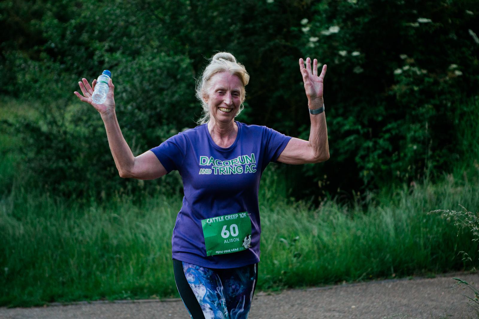 A woman in a purple running shirt with a race bib and "Dacorum and Tring AC" on it raises her arms in victory, holding a water bottle. She is running outdoors on a path, surrounded by lush green foliage, and is smiling.