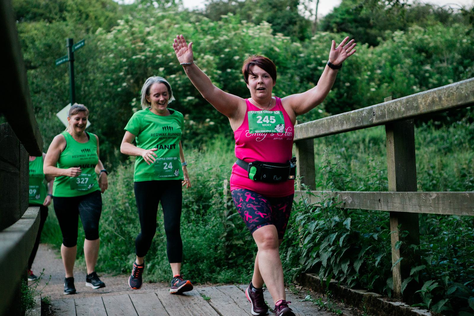 Three women running on a wooden path surrounded by green foliage. The woman in front, wearing a pink outfit, raises her arms in excitement, while the two women behind her, in green shirts, smile. Race bibs are visible on their shirts.
