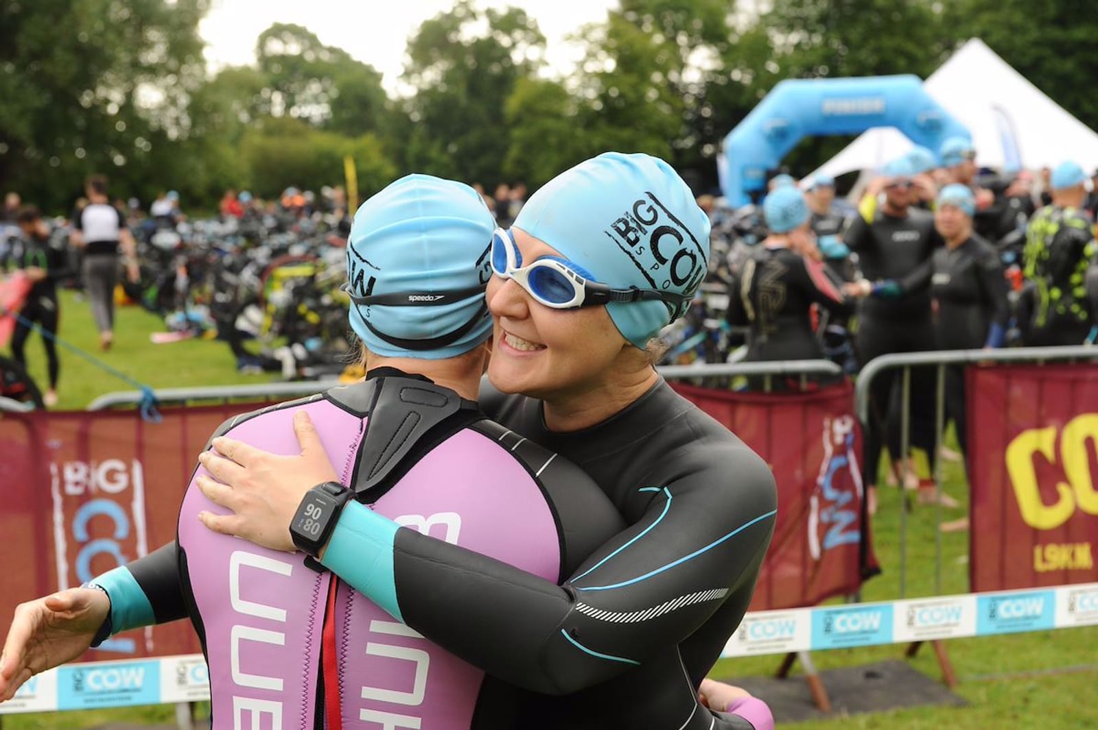 Two athletes in wetsuits and swim caps embrace near the starting section of a triathlon. One athlete wears a purple wetsuit and a smartwatch, while the other wears a black wetsuit and blue goggles. Other participants and spectators are visible in the background.