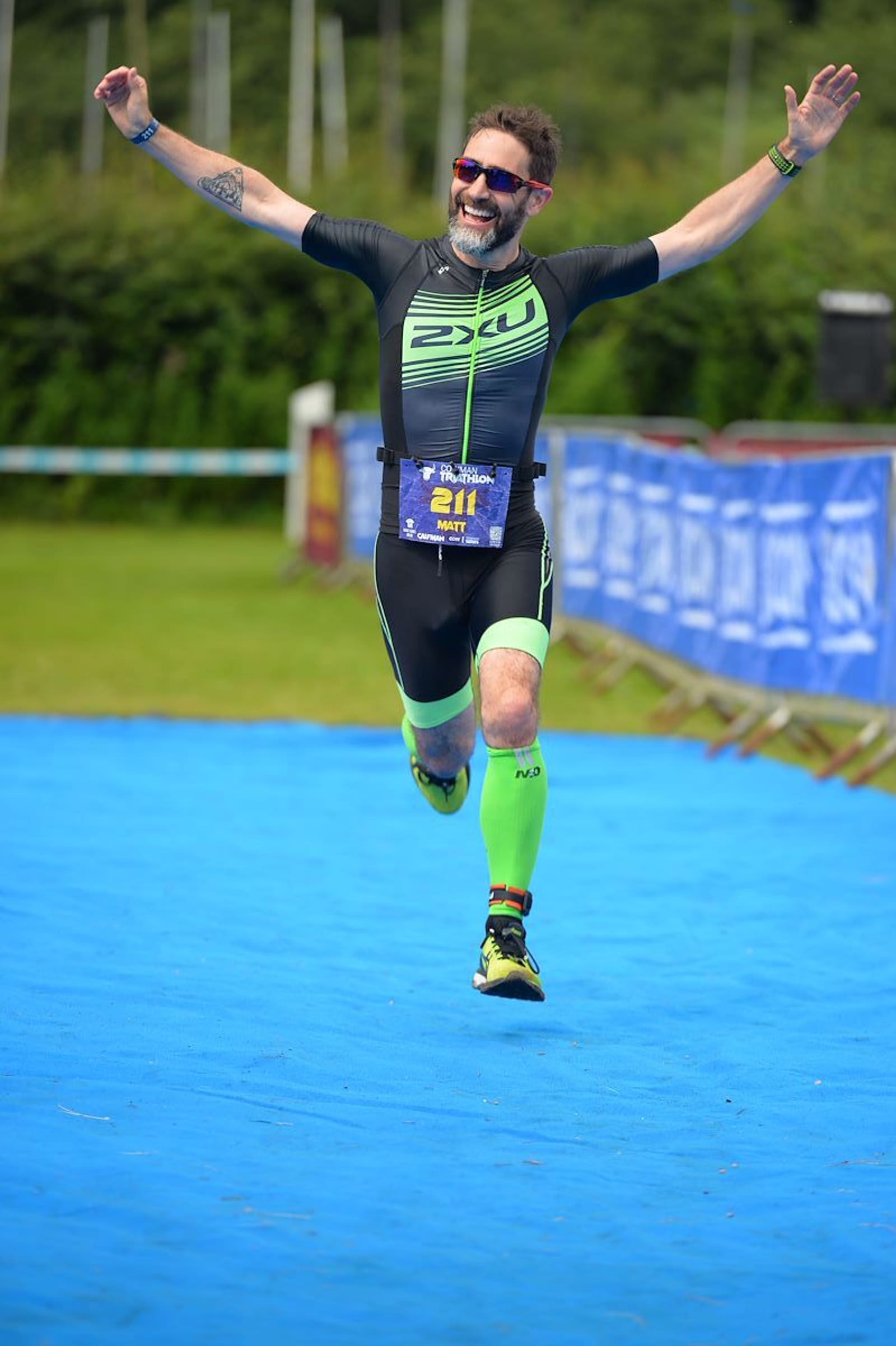 A male athlete wearing a black and green triathlon suit, sunglasses, and neon green calf sleeves runs joyfully towards the finish line with arms outstretched. The ground is covered with a blue mat, and the background features greenery and event banners.