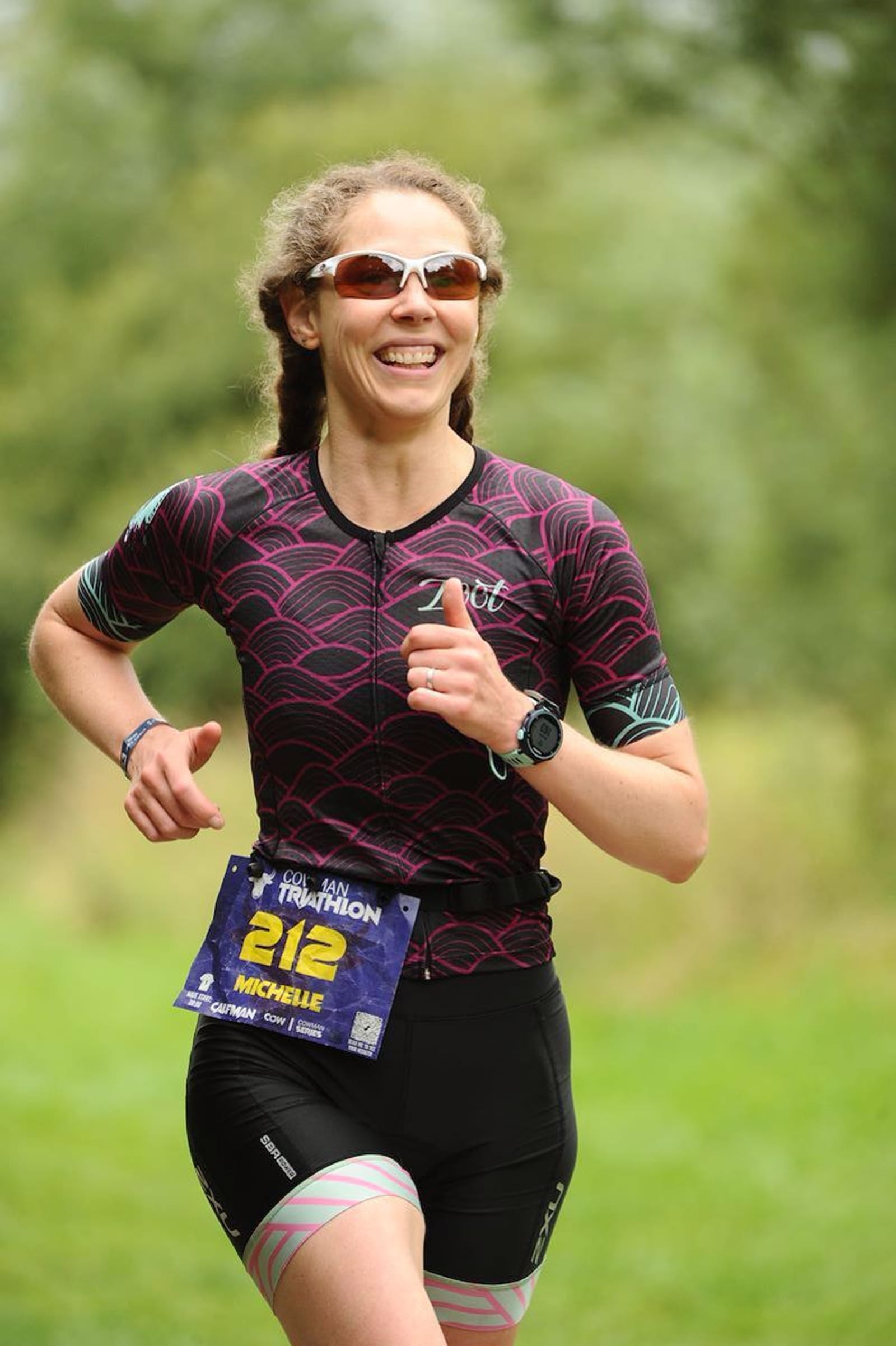A woman with curly hair, wearing sunglasses, a black and pink athletic outfit, and a race bib numbered 212, smiles as she runs outdoors on a grassy path. Trees and greenery are visible in the blurred background.