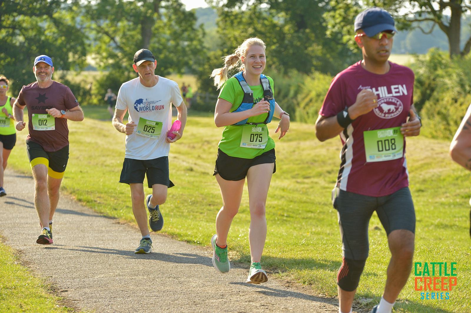 Four people are running on a path during a race event on a sunny day. They are wearing athletic clothing and race bibs. Lush green trees and grass are in the background. The event logo "Cattle Creep Series" is in the bottom right corner.