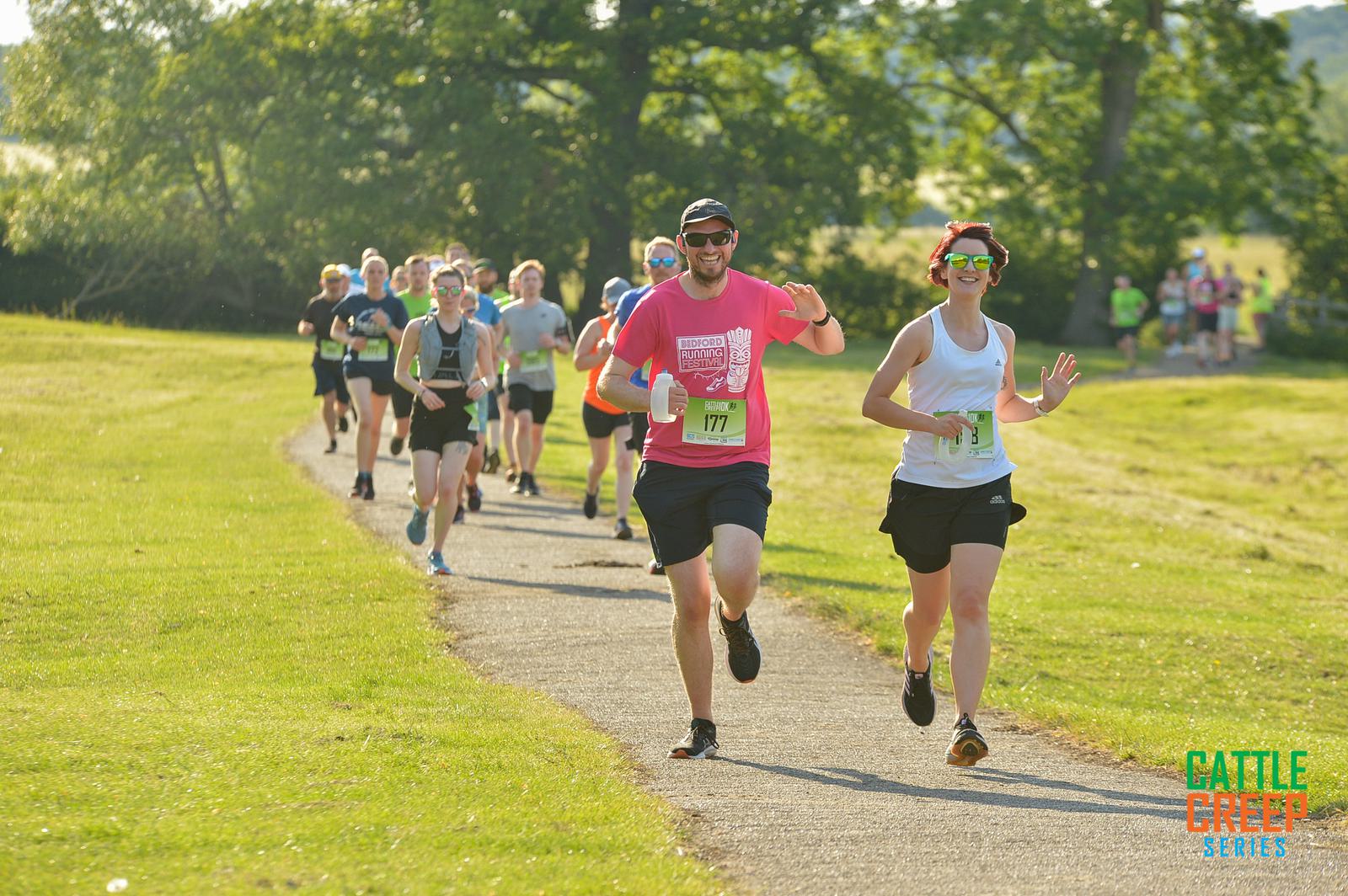 A group of runners, wearing various athletic outfits and bibs, run on a sunny park trail surrounded by lush greenery. The participants appear joyful, with some waving and smiling at the camera. "Cattle CreeK Series" is visible in the corner.