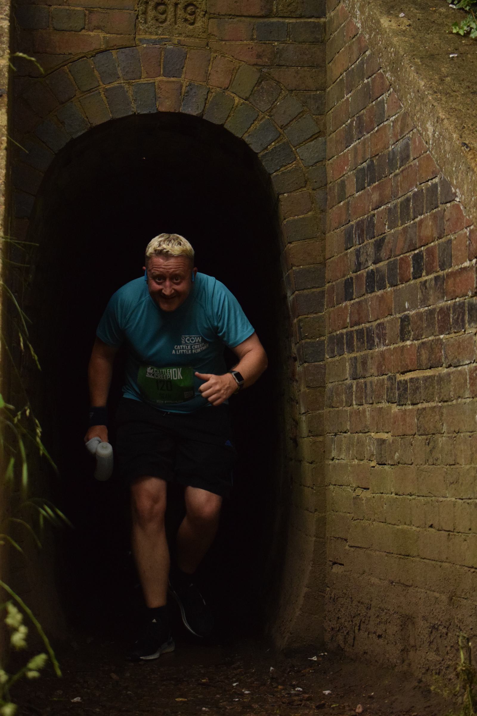 A man in a blue shirt and black shorts emerges from a dark brick tunnel, smiling and holding a water bottle in his right hand. The tunnel is surrounded by greenery.