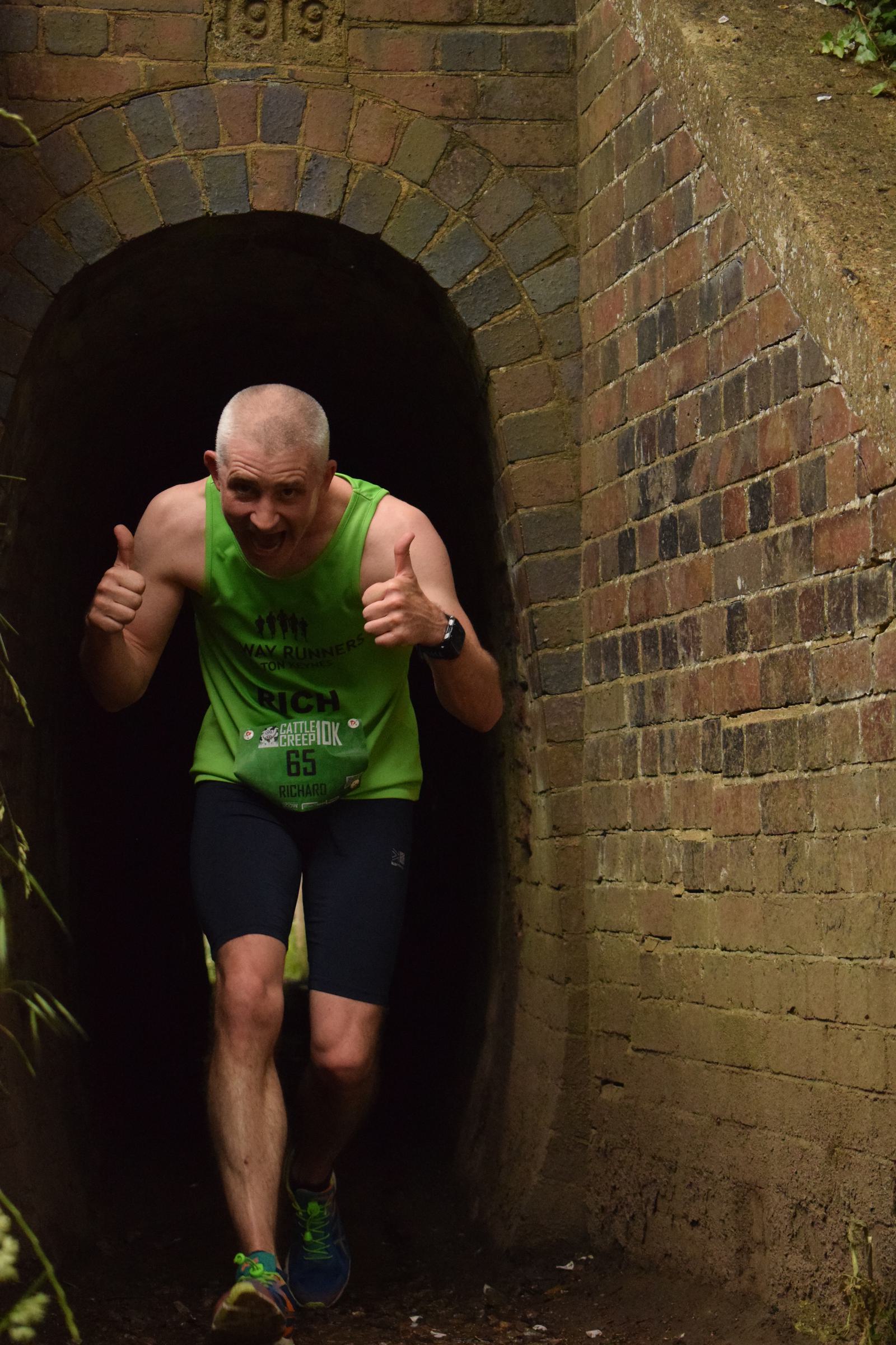 A man in a green tank top and black shorts runs through a narrow brick tunnel. He is smiling and giving a thumbs-up with both hands.
