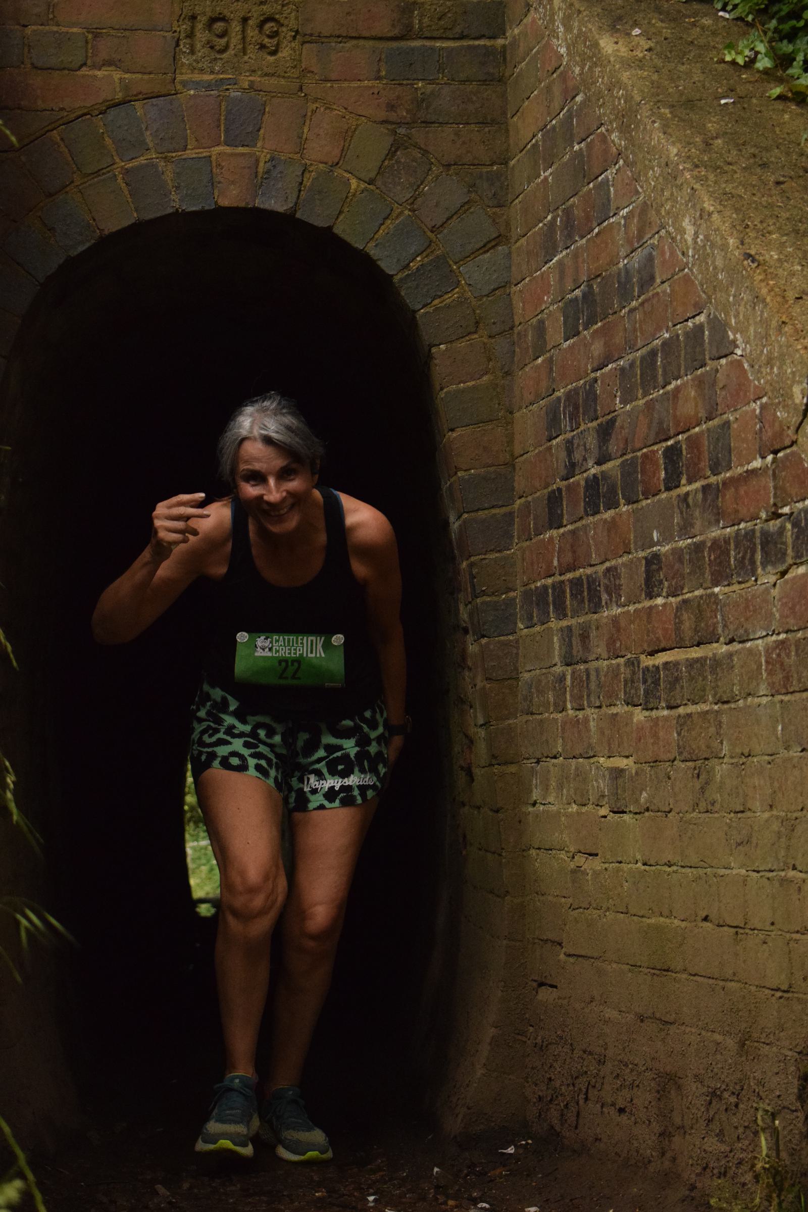 A person with short gray hair is smiling while running through a brick tunnel. They are wearing a sleeveless black top and green leopard-print shorts. The year "1919" is engraved above the tunnel.