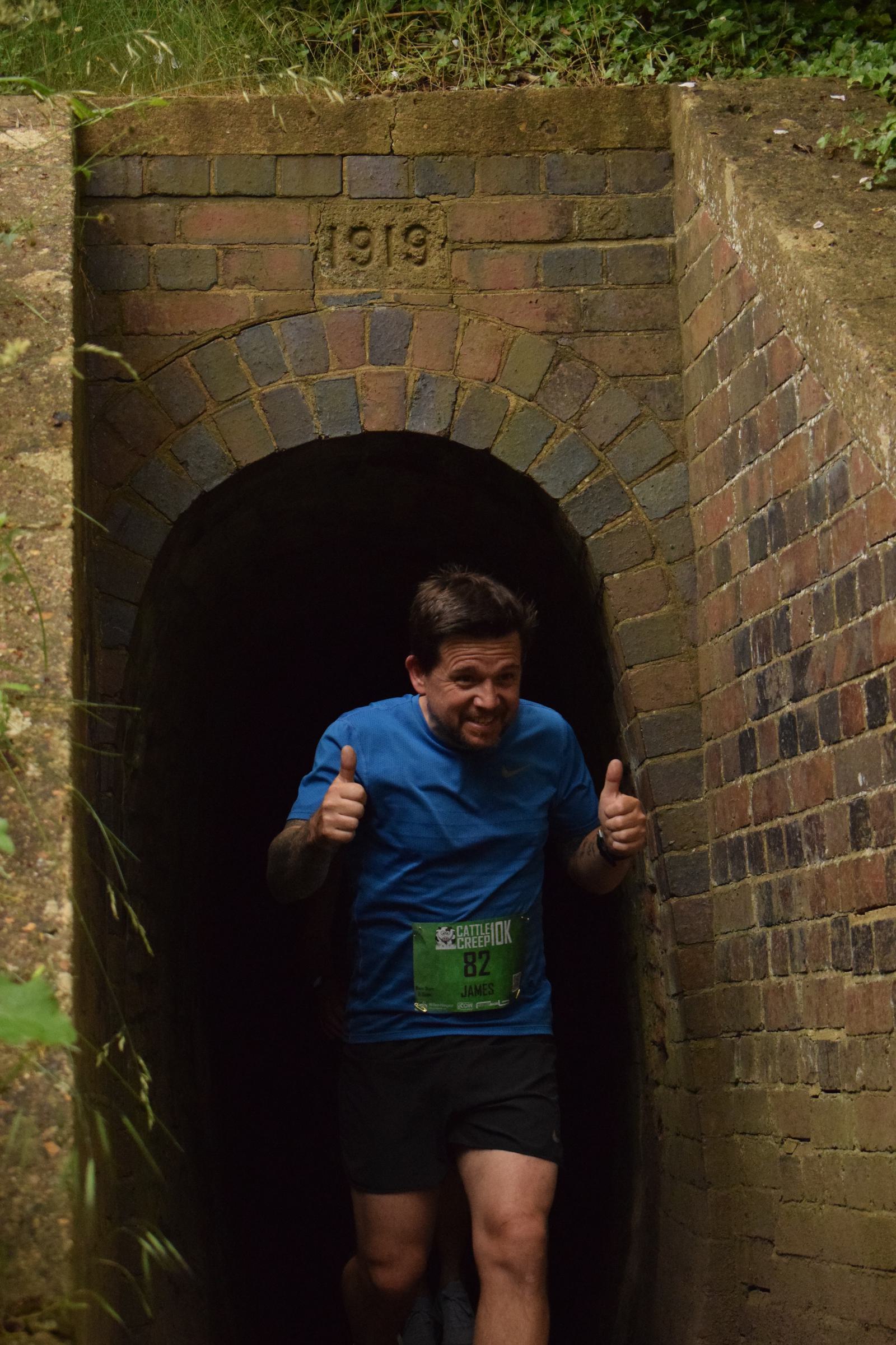 A man in a blue shirt is jogging through a brick tunnel with "1919" etched above the entrance. He is giving two thumbs up and wearing a race bib labeled "B2." Green foliage surrounds the tunnel.