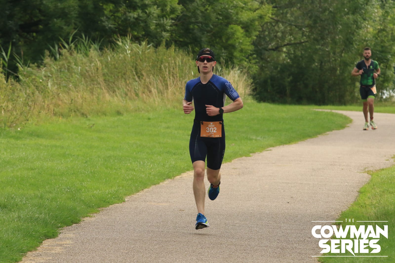 A man wearing a blue athletic suit and sunglasses runs on a paved path during a race, with another runner in the background. The event logo "The Cowman Series" is visible in the bottom right corner.