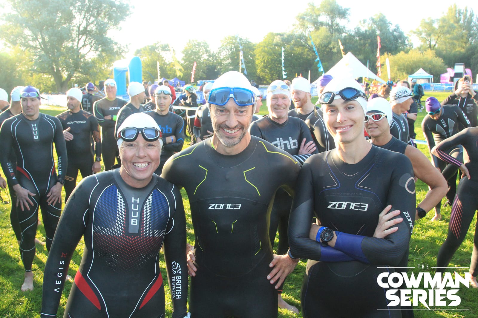 A group of smiling triathletes in wetsuits and swim caps stand together outdoors before a race, with more participants and trees visible in the background. "The Cowman Series" logo is in the bottom right corner.