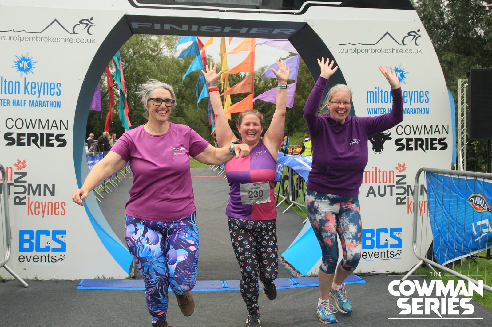 Three women in colorful athletic wear joyfully raise their arms as they cross the finish line at a running event, smiling beneath a large finish arch with "The Cowman Series" branding in the background.